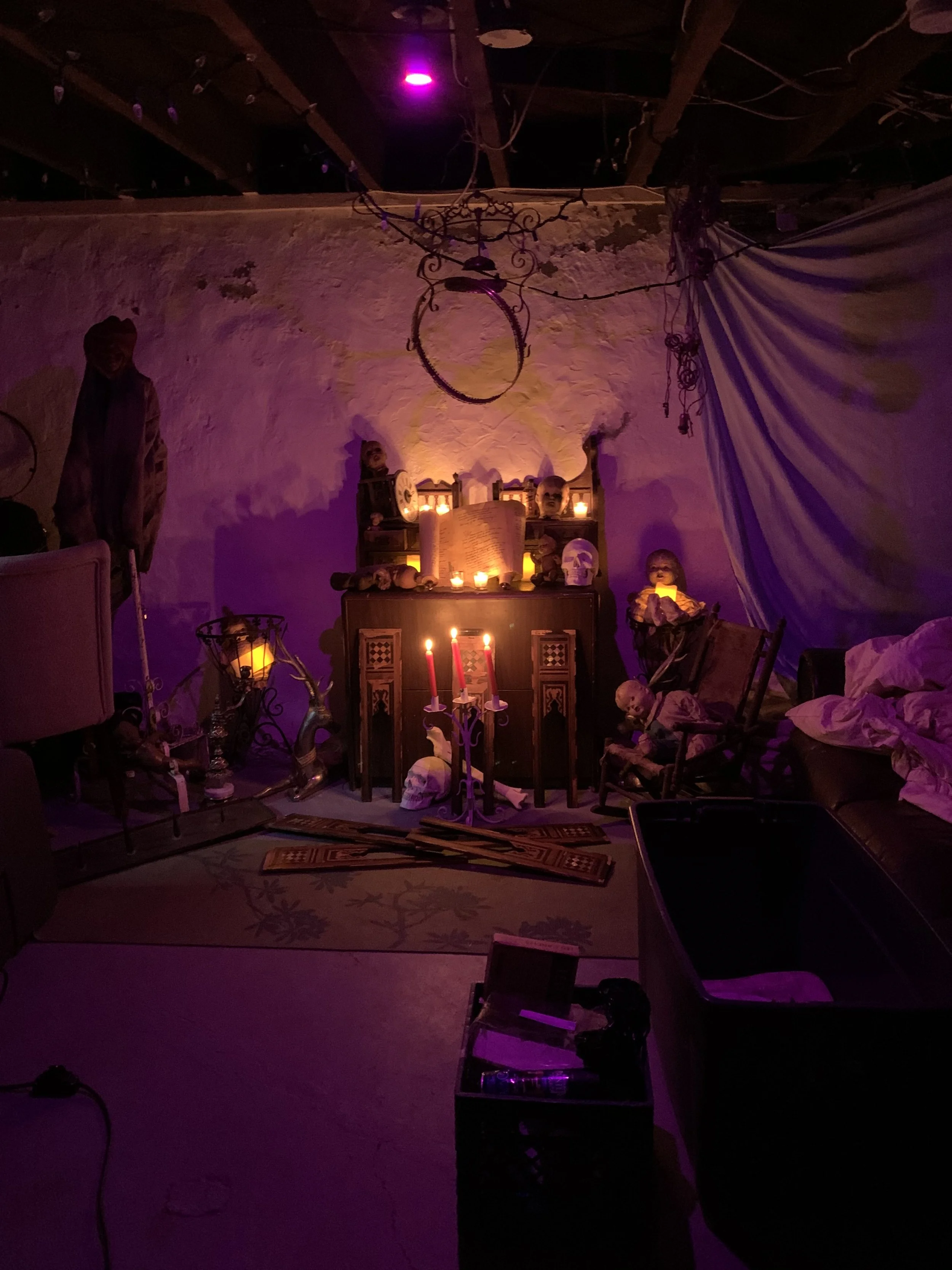 A dimly lit room decorated with Halloween or spooky-themed items, including candles, skulls, and skeletons. The room has a rustic, vintage vibe with a mix of eerie decorations and furniture such as chairs, tables, and a cabinet, creating an eerie, ha