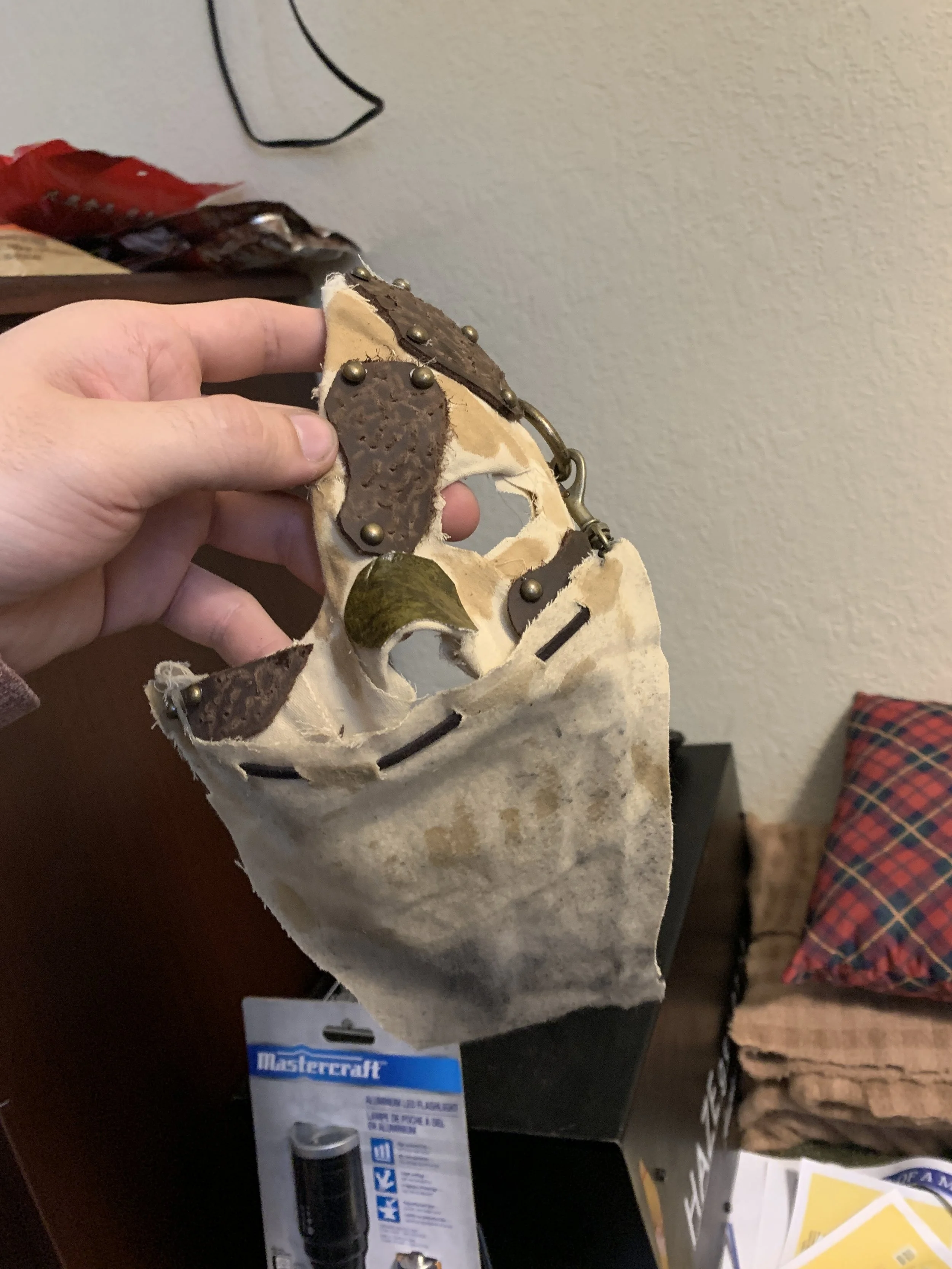 A person's hand holding a beige and brown decorative item with metal studs and leather accents, standing over a DIY project with an EGO power tool package and a stack of plaid fabric on a table.