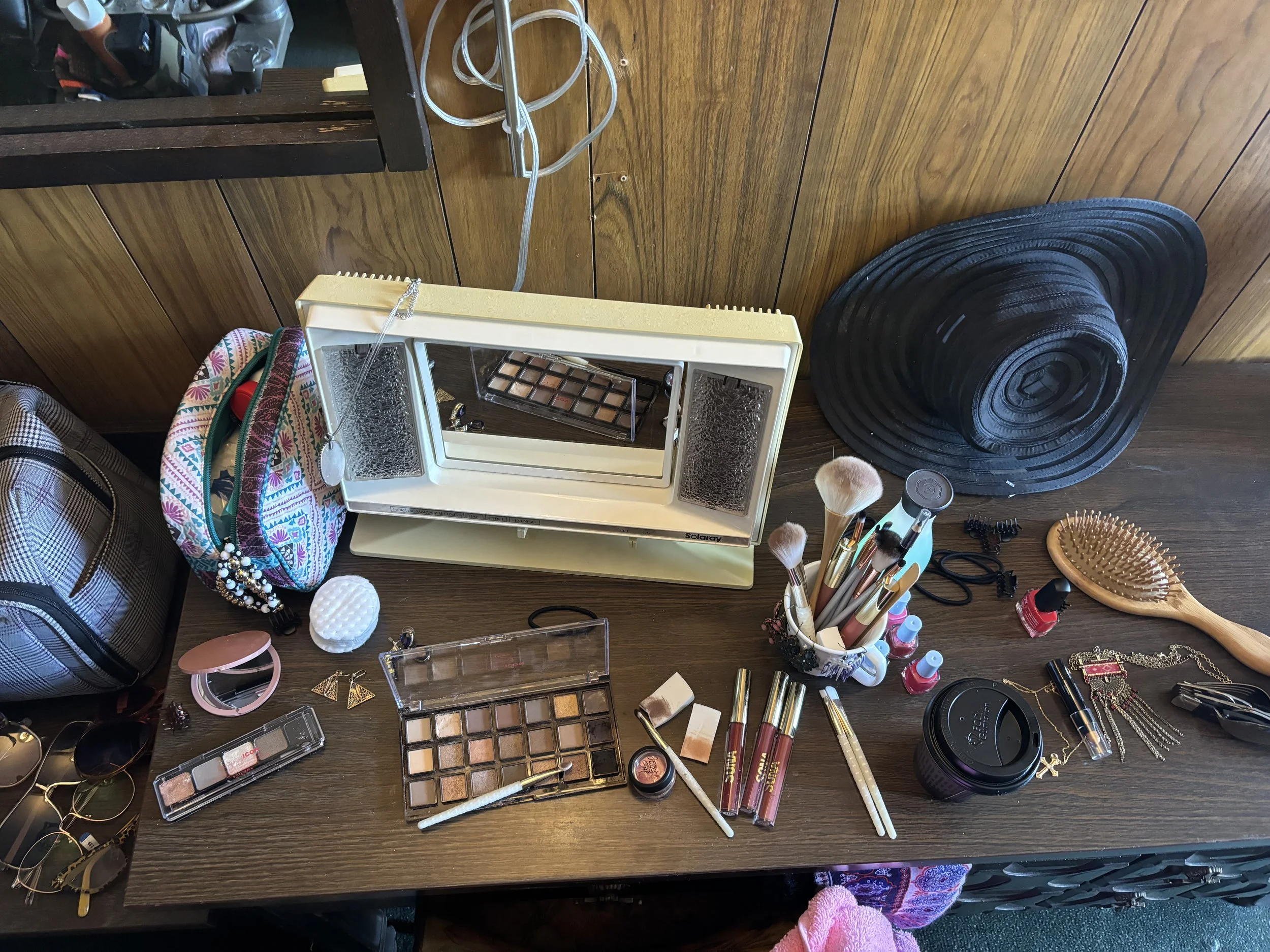 A cluttered wooden table with various makeup and jewelry items, including an eyeshadow palette, makeup brushes, lipstick, sunglasses, perfume, jewelry, and a patterned bag.