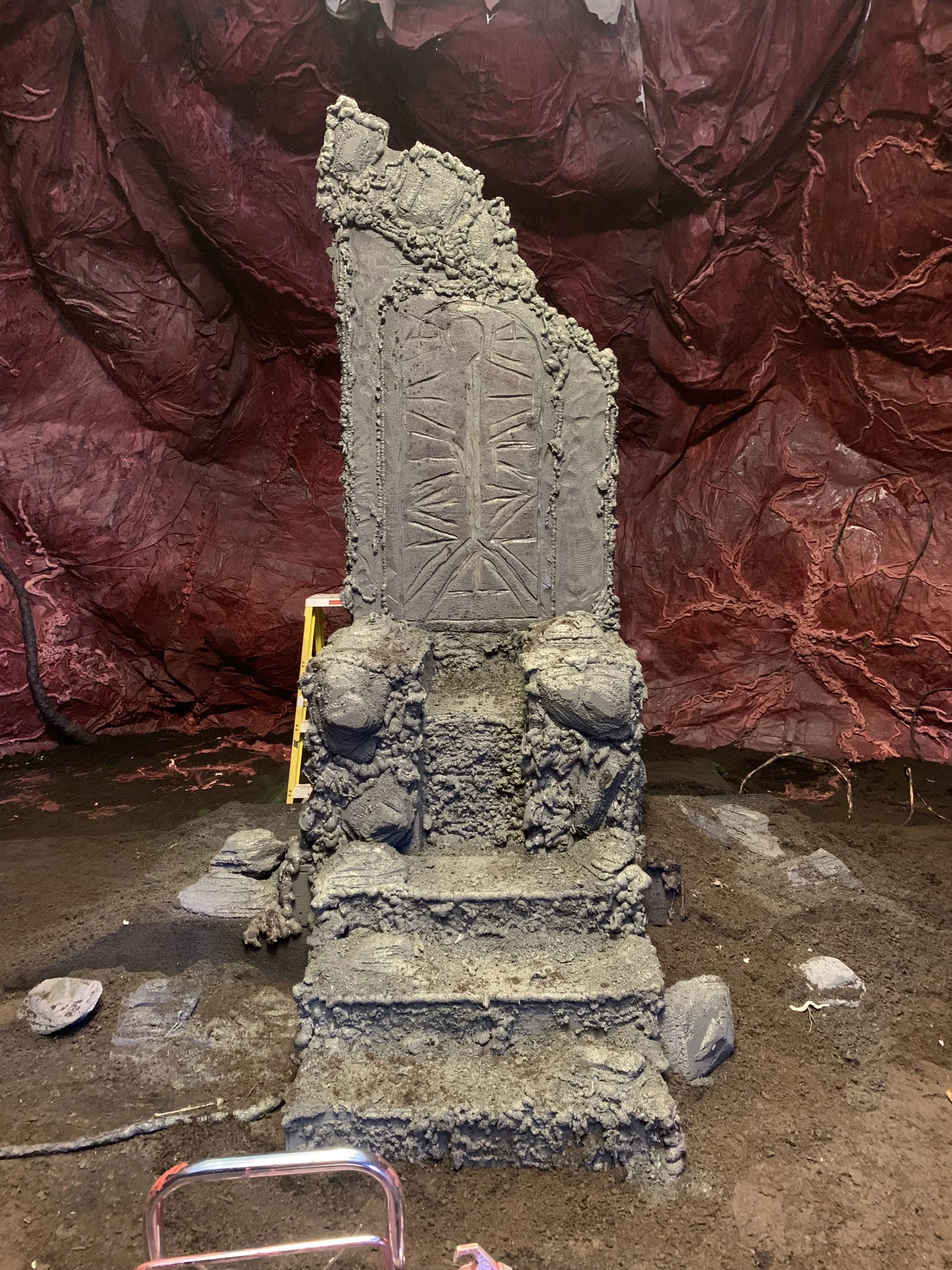 Ancient stone stele with carvings, situated in an underground cave with reddish rock walls, scattered rocks, and dirt floor.