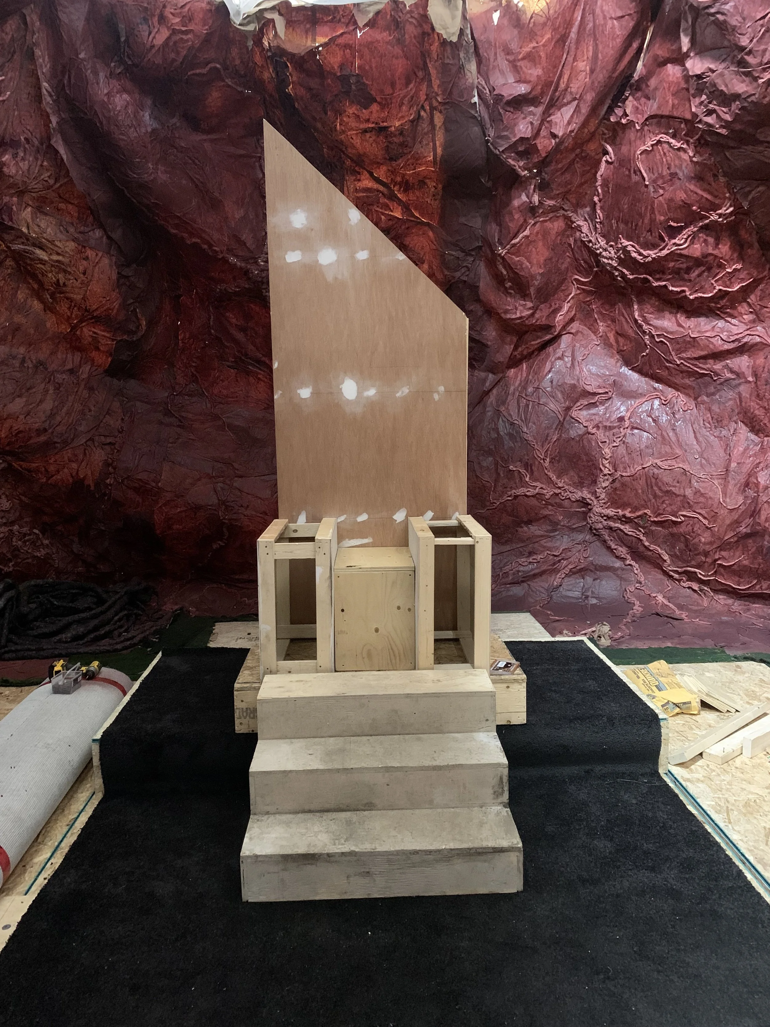 A partially assembled wooden structure on a platform with steps in front, set against a large red rock formation backdrop. Construction tools and materials are visible around the workspace.