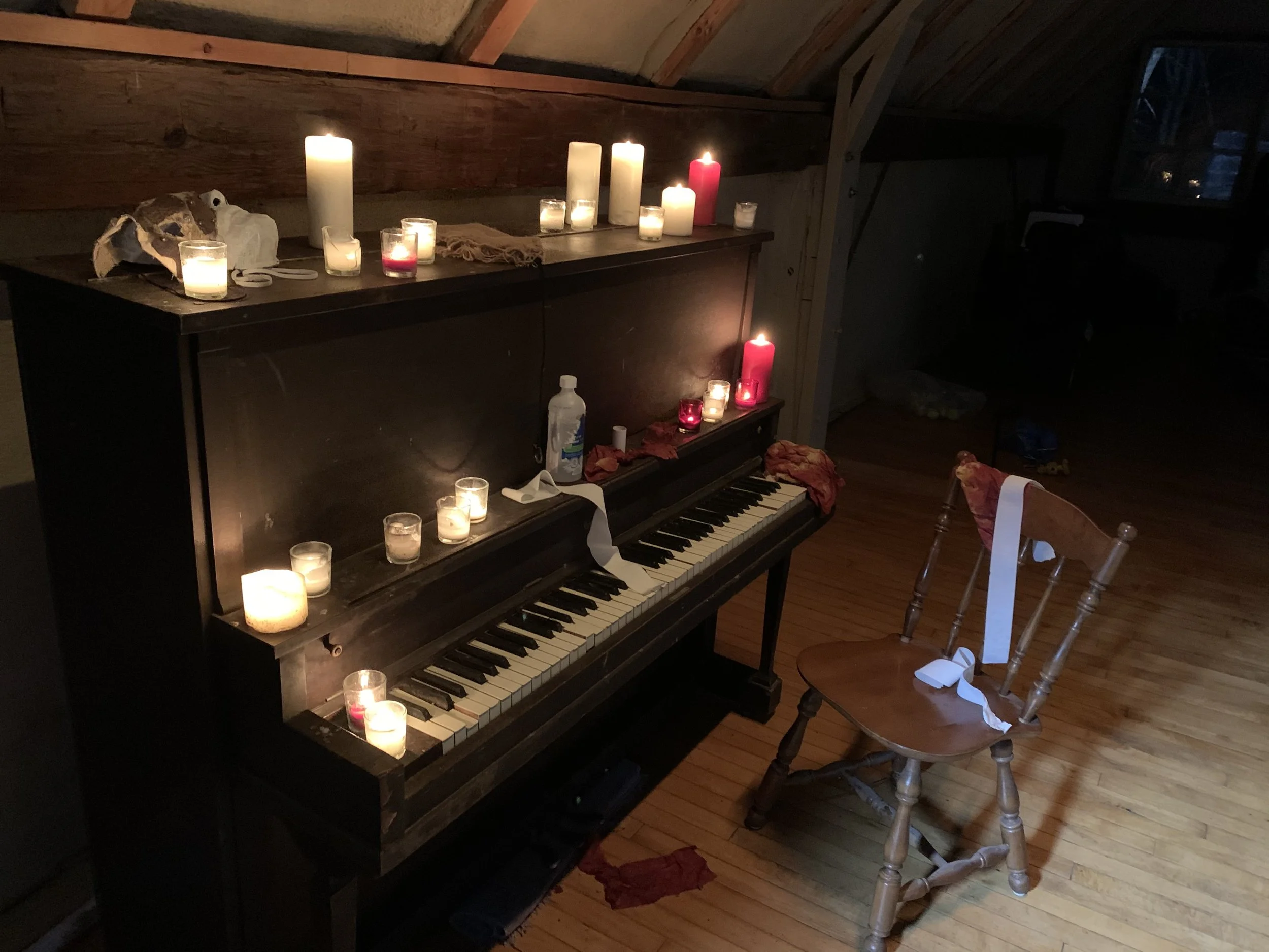 A black upright piano with various lit candles on top. There is a wooden chair with a red cloth and a white ribbon tied around the backrest positioned in front of the piano. The room has wood-paneled walls and hardwood floors.