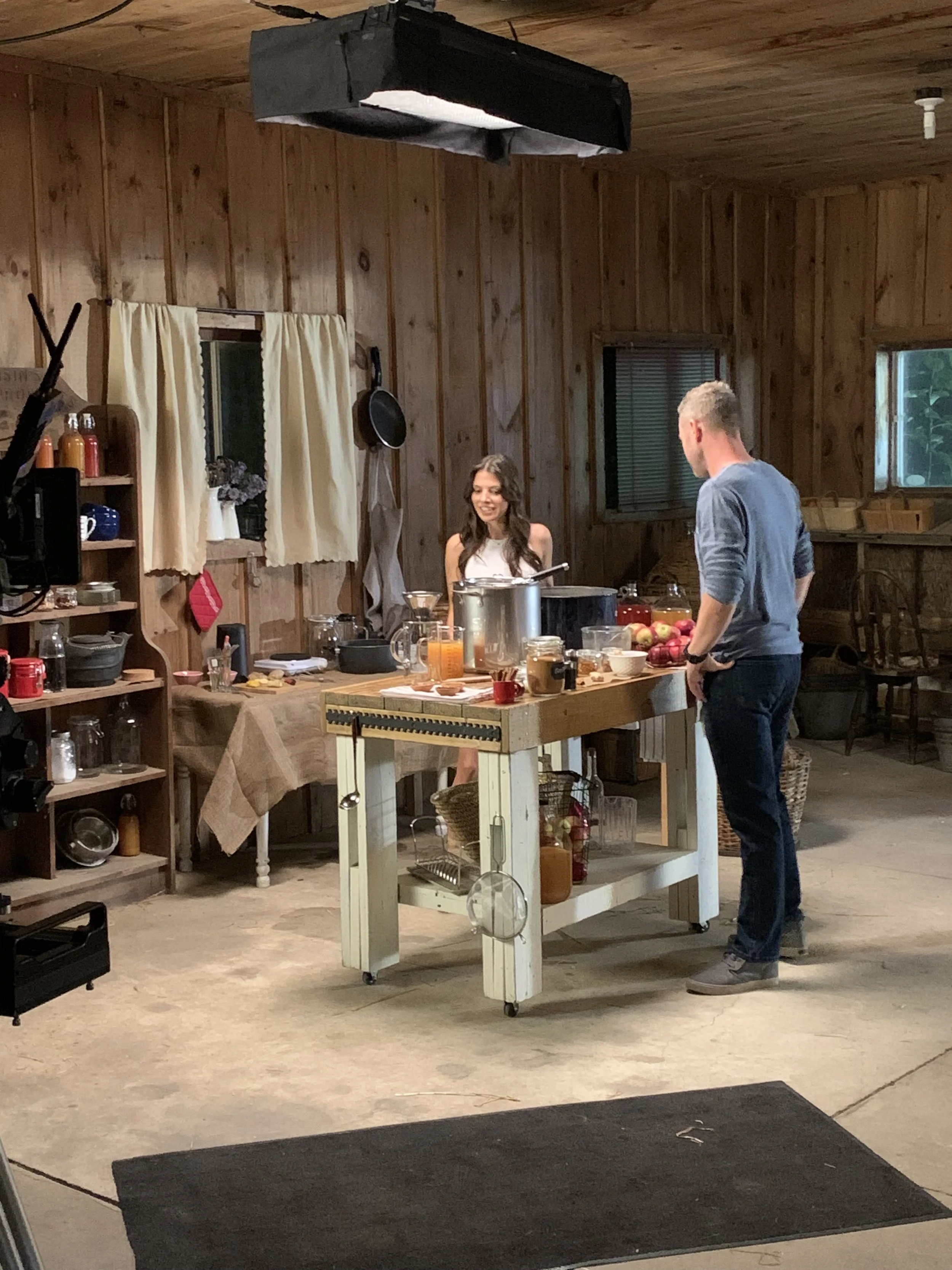 Two people in a rustic kitchen with wood-paneled walls. The woman, wearing a white top, is smiling and standing behind a kitchen island with various kitchen tools and ingredients. The man, wearing a blue long-sleeve shirt and jeans, is facing her. Th