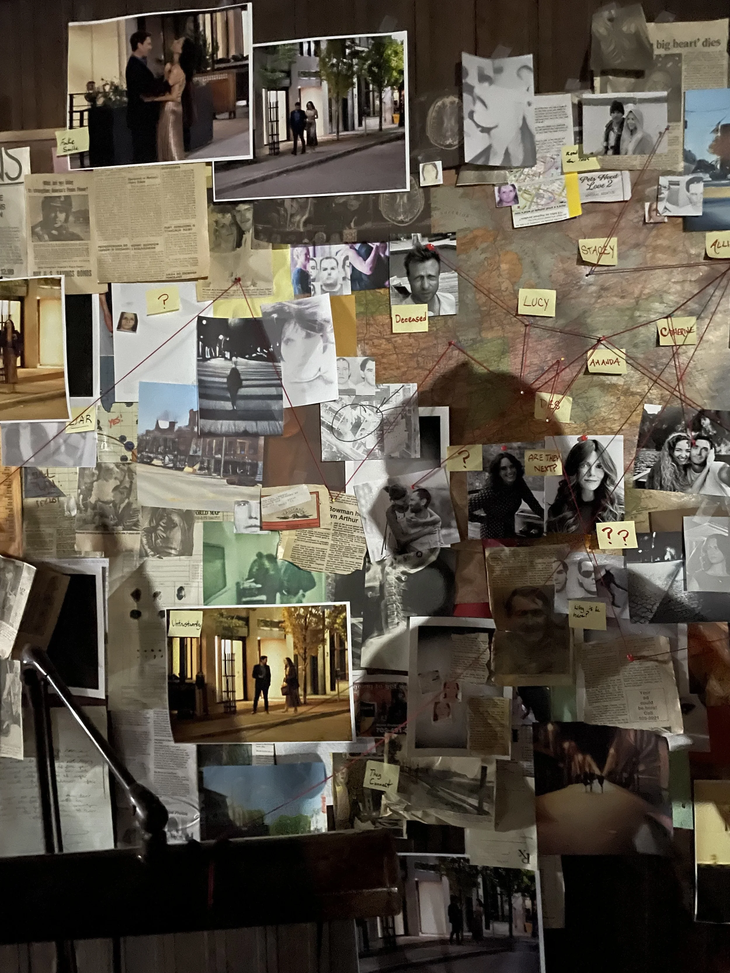 A wall decorated with photos, newspaper articles, notes, and string connecting various items. The photos include people, some in black and white and others in color, with scenes of city streets and indoor settings. Post-it notes with handwritten labe