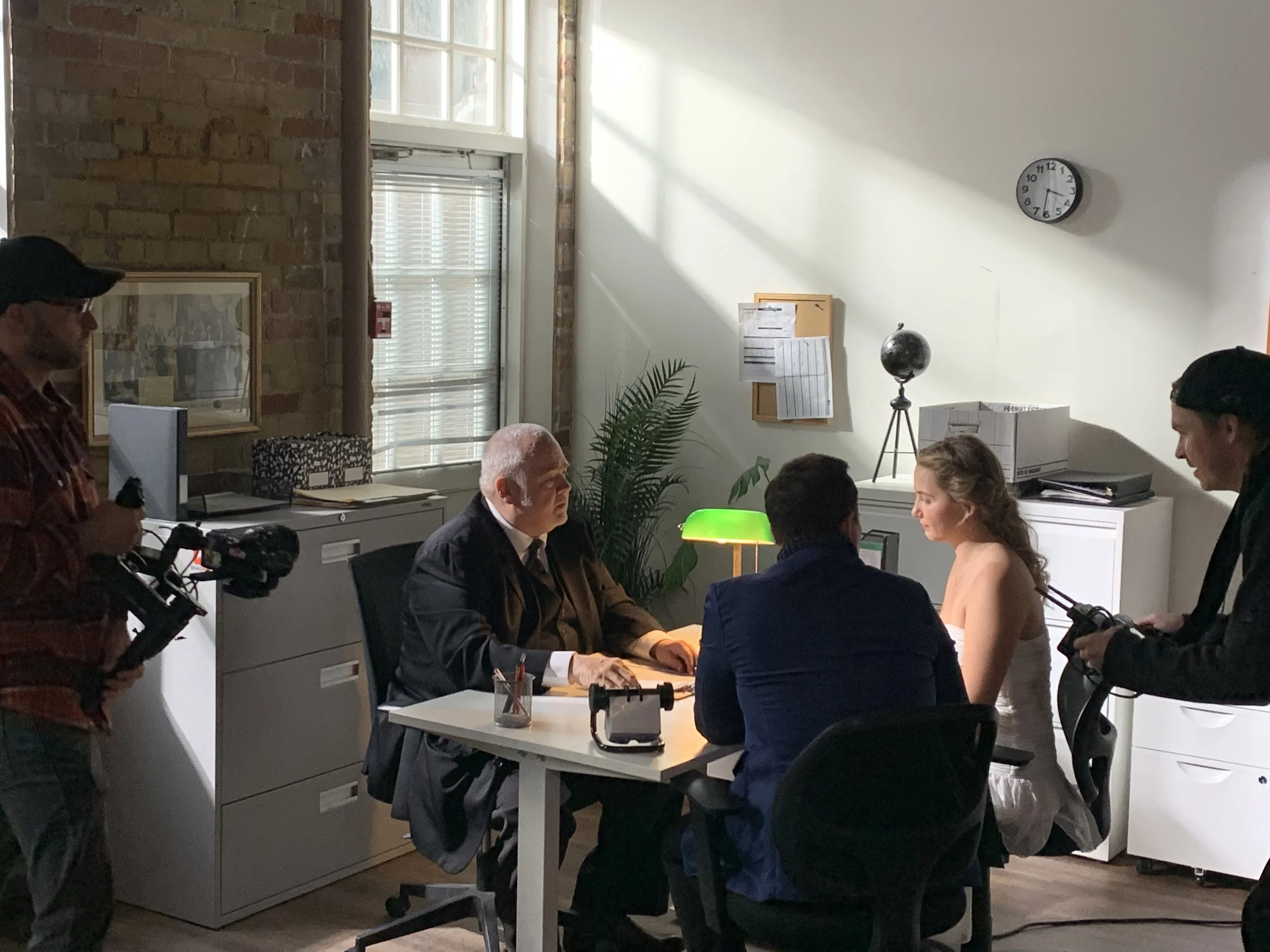 A group of people in a professional setting, some seated at a desk while others stand around, engaged in conversation. The room has a brick wall, large window, and office furniture, indicating an office or meeting room.
