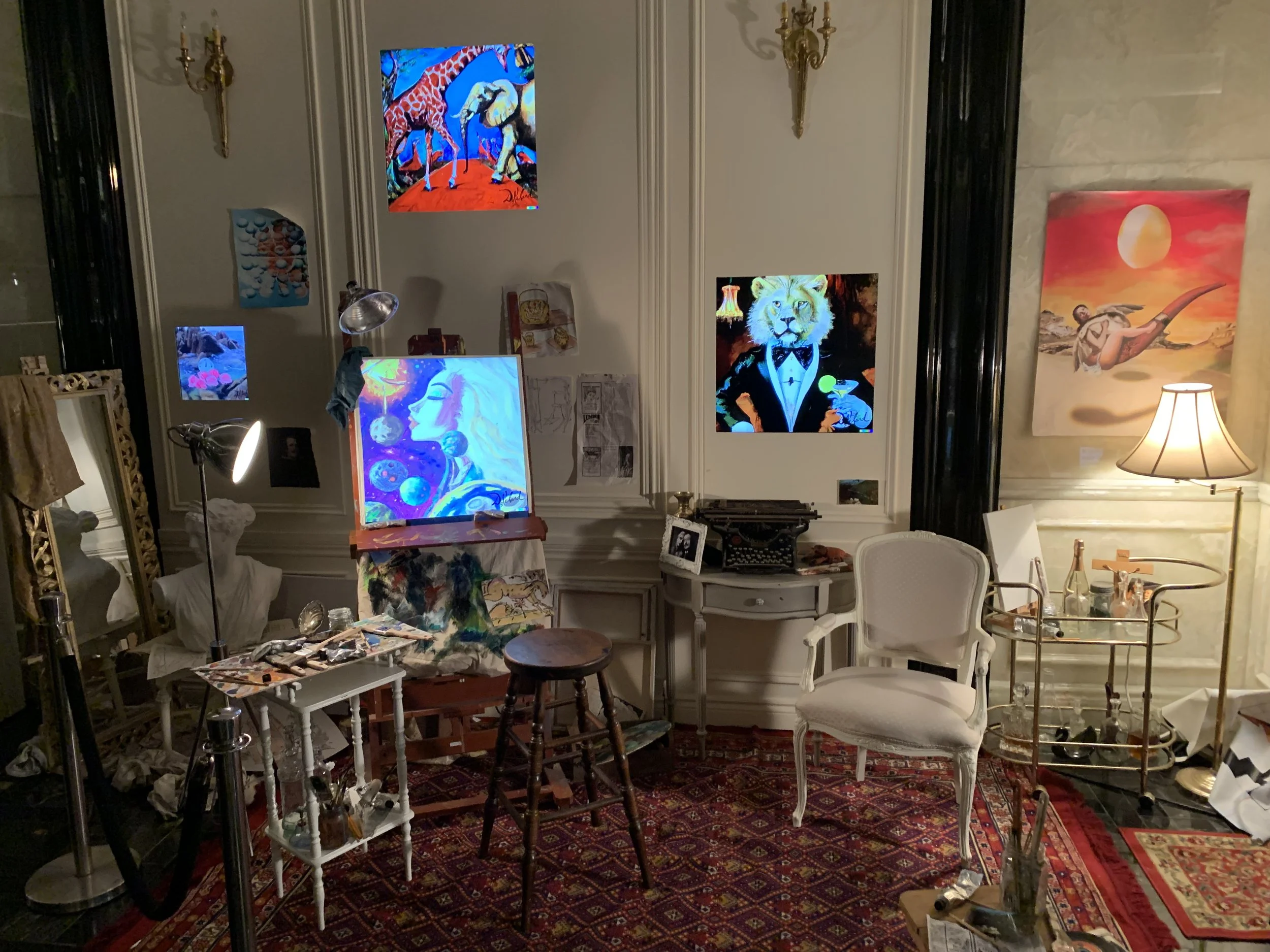 The image shows an artist's studio with various paintings displayed on the walls, including colorful, artistic depictions of animals and abstract designs. The studio is cluttered with art supplies, easels, chairs, and lighting equipment, and is decor
