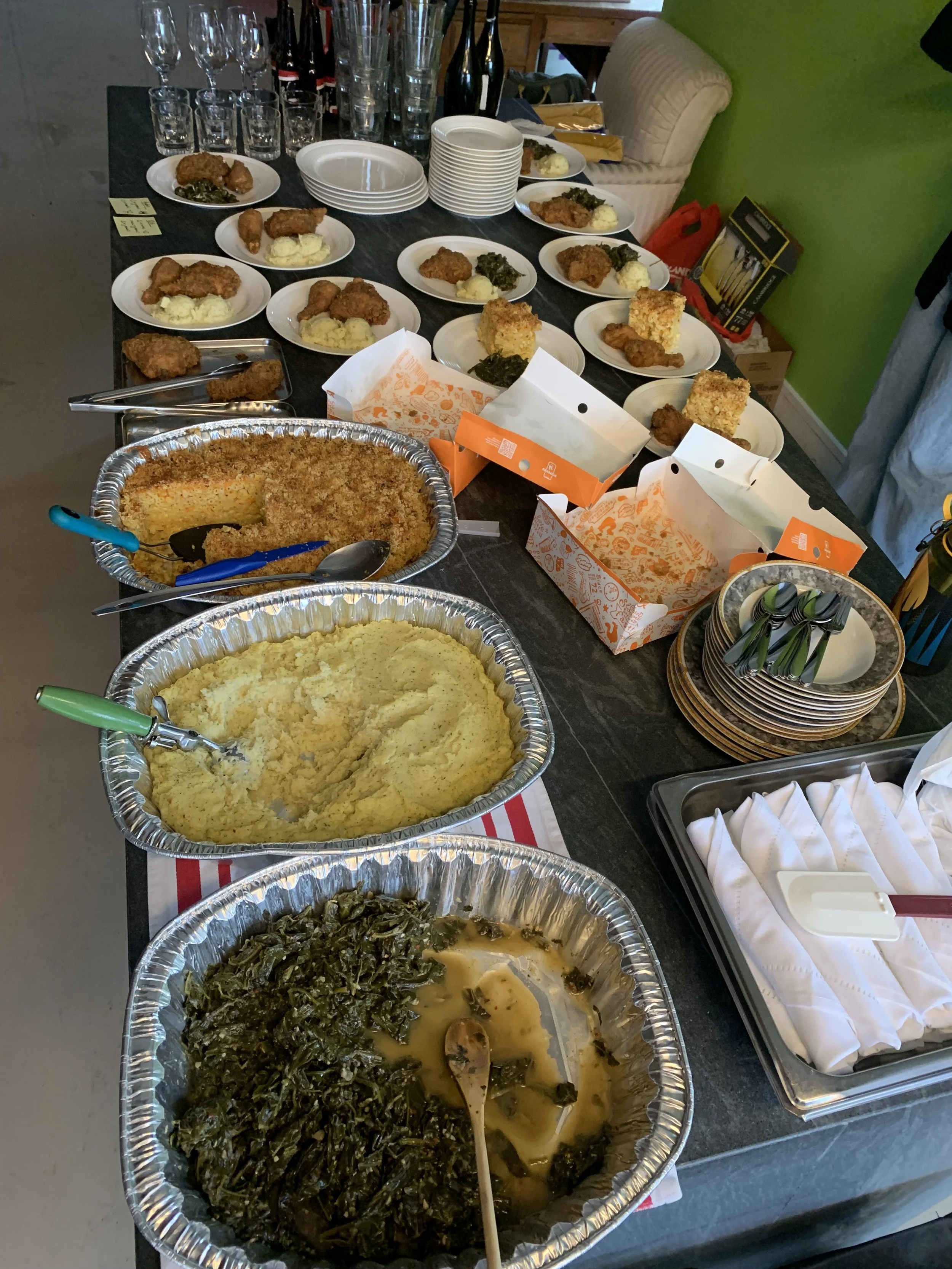 A buffet table with plates of fried chicken, mashed potatoes with gravy, green leafy vegetables, baked casserole, and other dishes. There are empty glasses and utensils, with some packaging for food to go on the table.