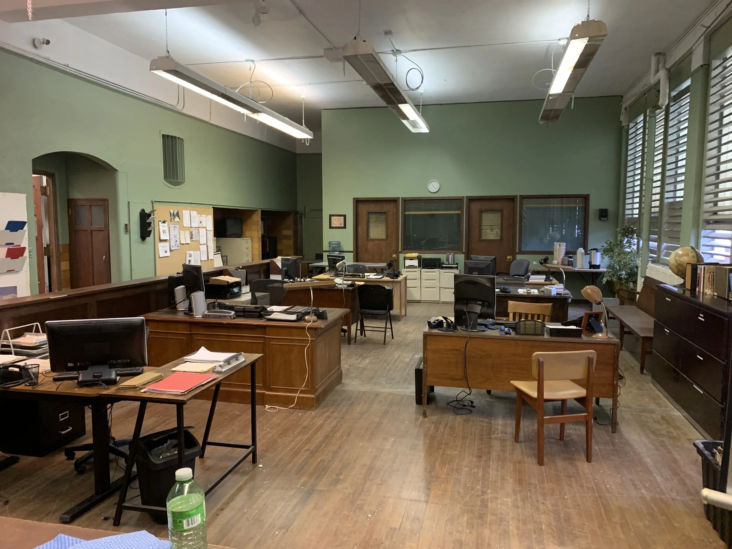An office room with green painted walls, several desks, chairs, computers, and office supplies scattered around. The room appears cluttered and is illuminated by fluorescent lights hanging from the ceiling. There are large windows with blinds on the 