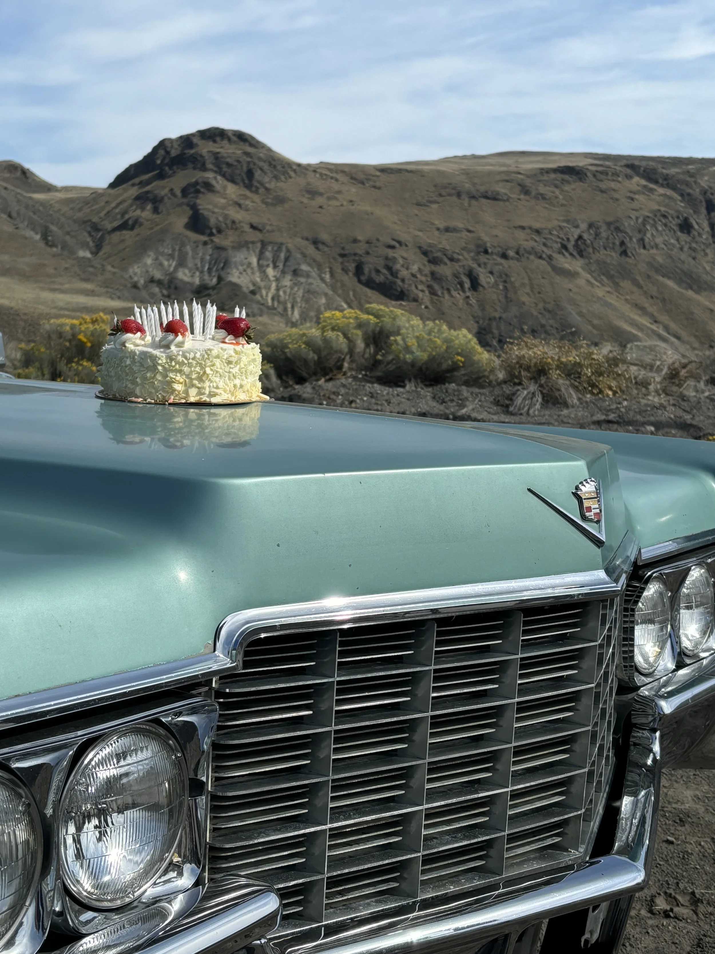 A vintage turquoise Cadillac car with a cake on its hood, featuring strawberries and candles, parked in a desert landscape with rocky hills and sparse vegetation.