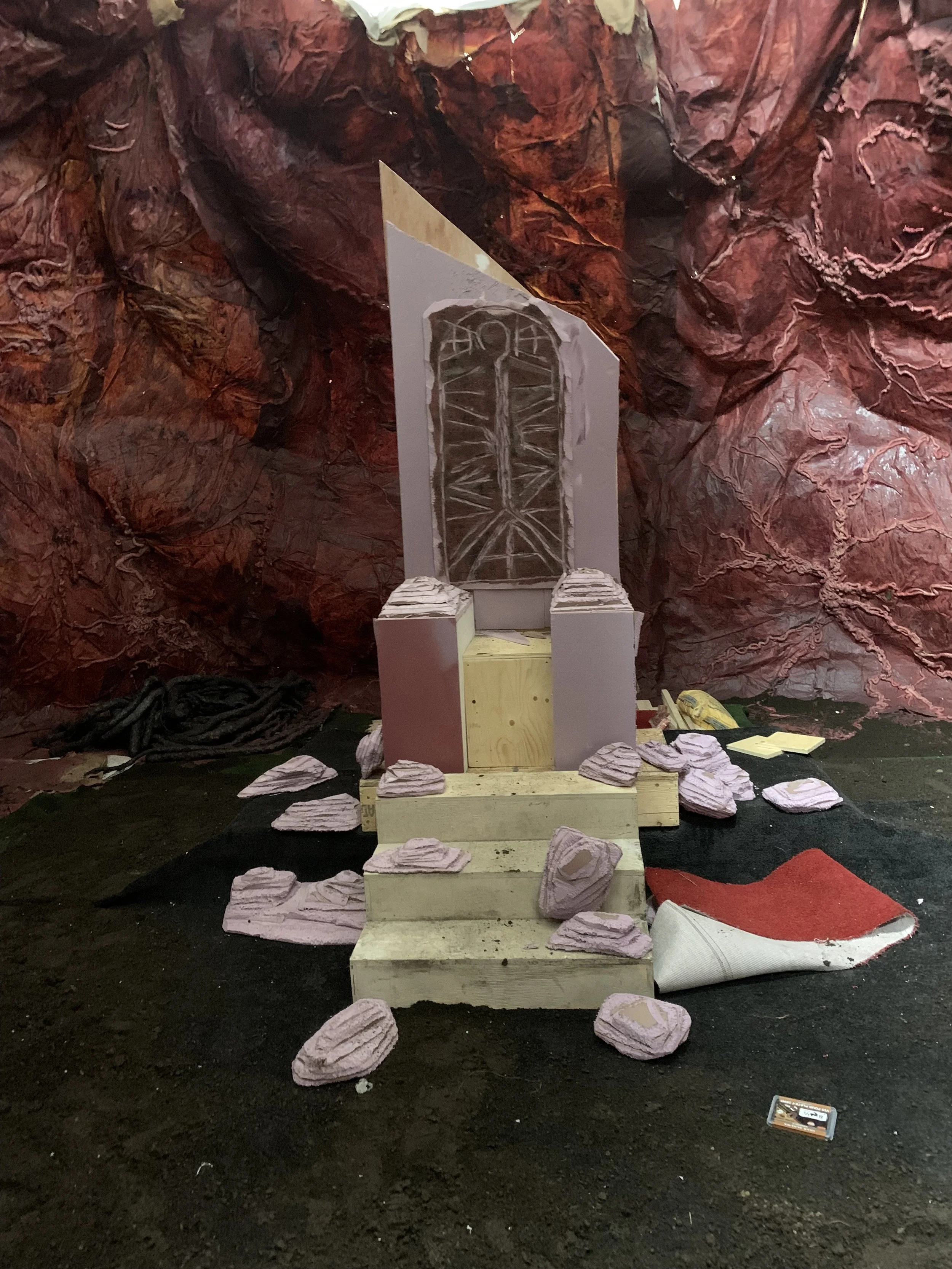 An art installation or stage set with a backdrop of red-brown crumpled paper or fabric. In the center, a structure resembling a white archway with the word 'FAITH' written above it, constructed from white and yellow wood. Surrounding the structure ar