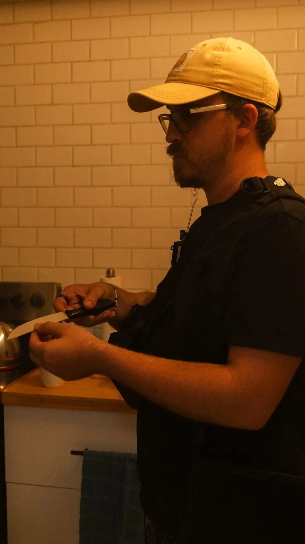 A man wearing glasses, a beige cap, and a black shirt is using a knife to cut something over a piece of paper, standing in front of a brick wall in a kitchen.