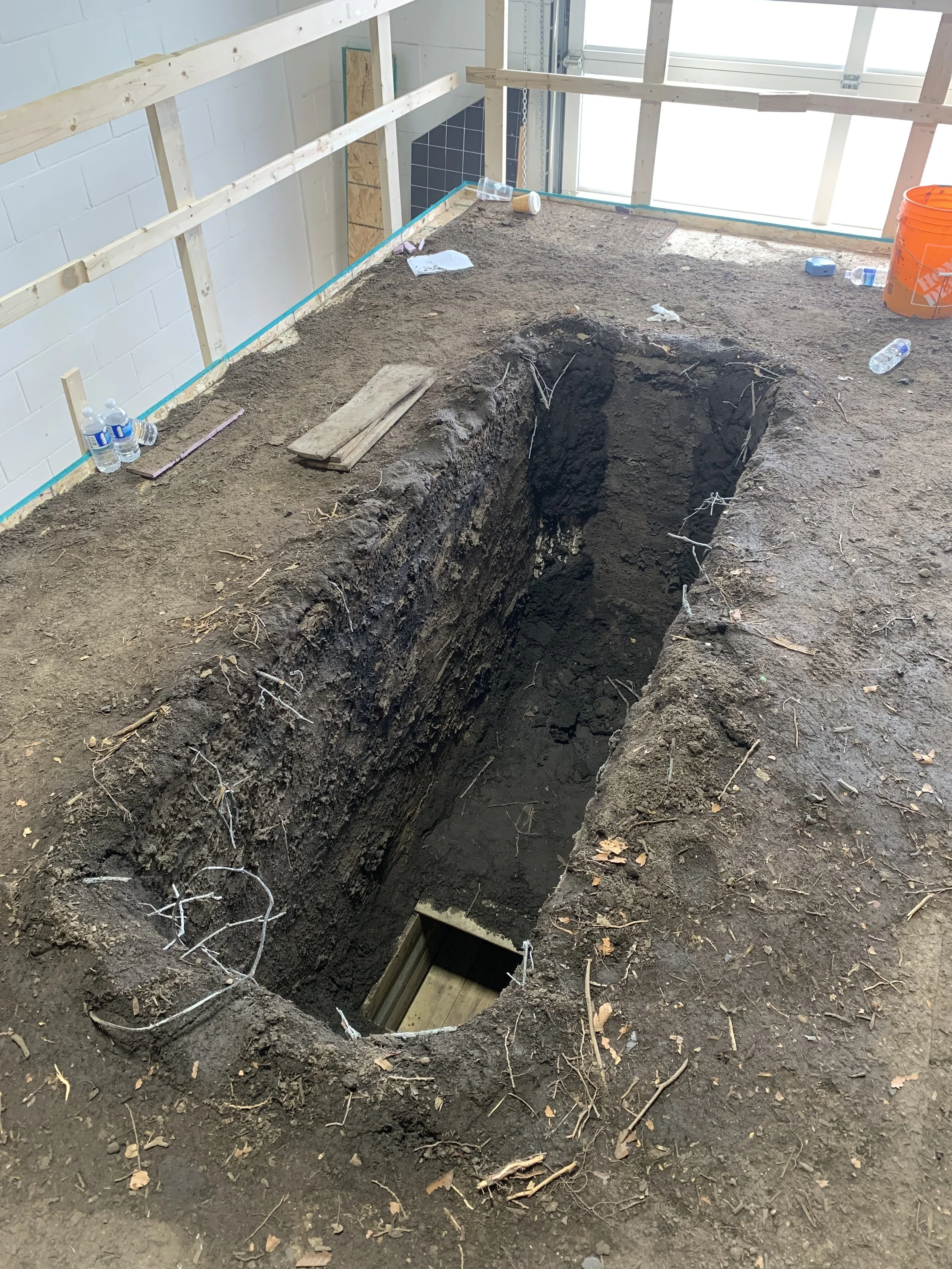 A construction site with a large, deep trench in the dirt floor, surrounded by wooden framing and construction materials, including water bottles, a bucket, and tools.