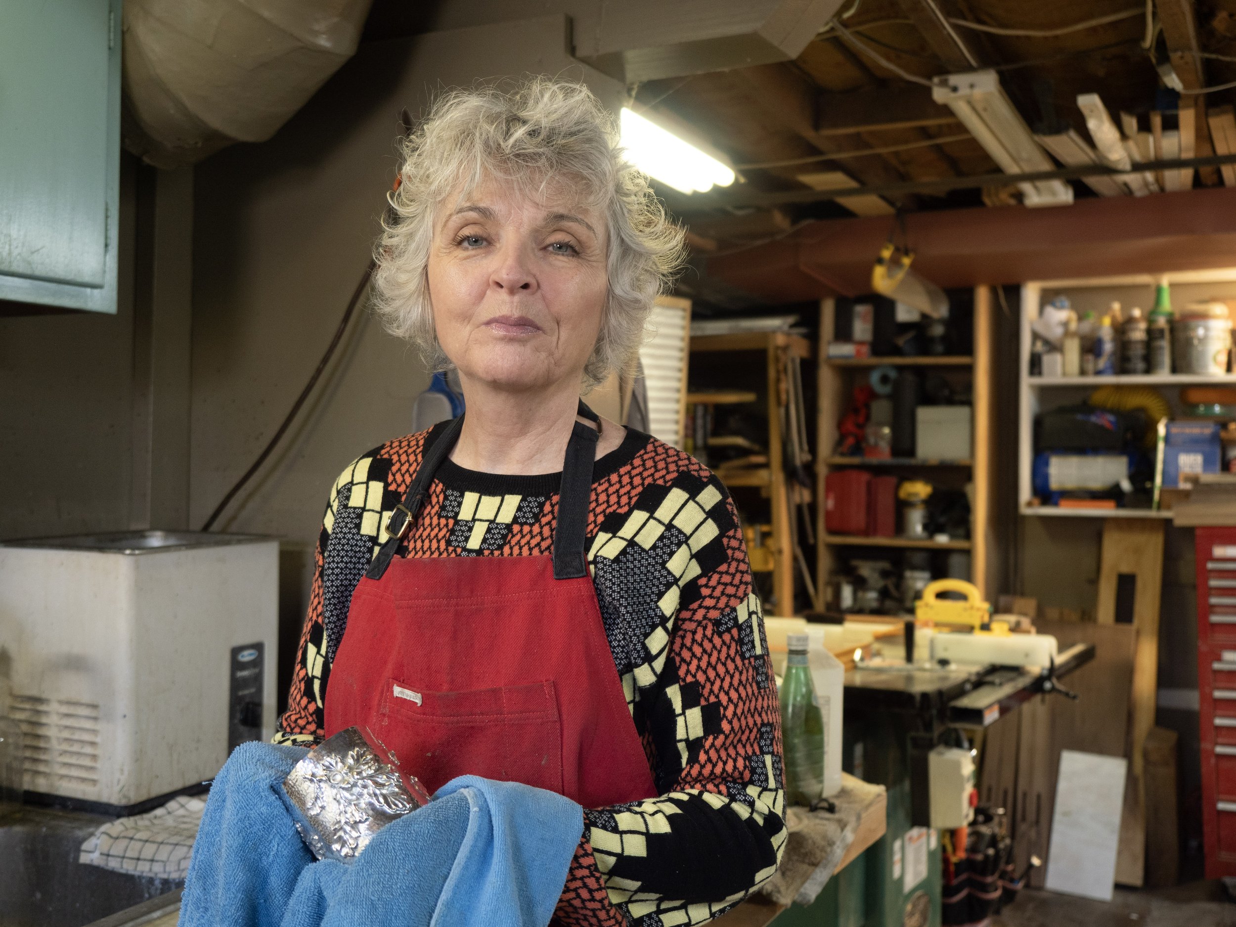 A woman with short, curly, gray hair wearing a colorful patterned sweater and a red apron, standing in a workshop or garage.