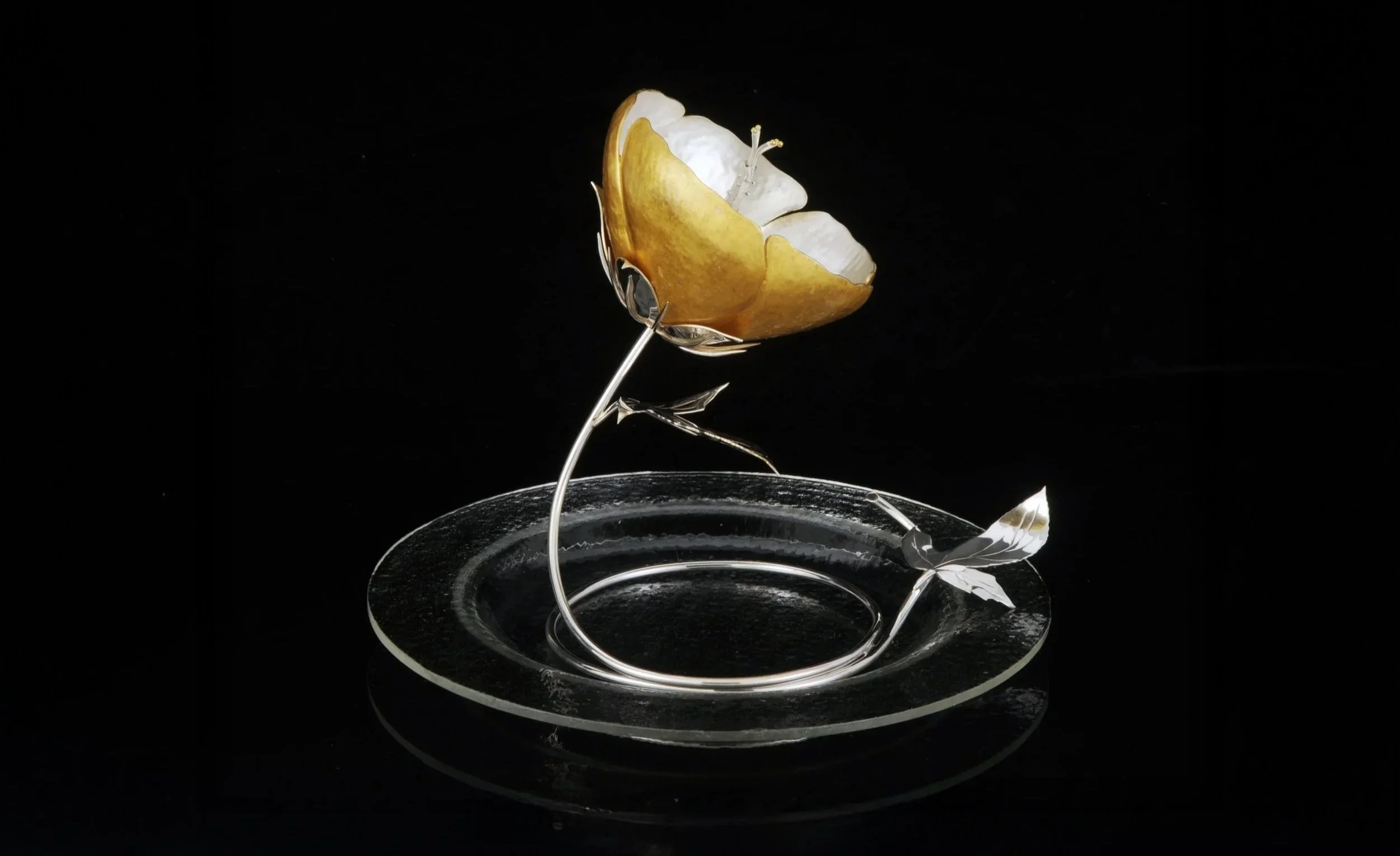 A gold-colored butterfly sculpture with silver accents, positioned on a clear glass or acrylic base against a black background.
