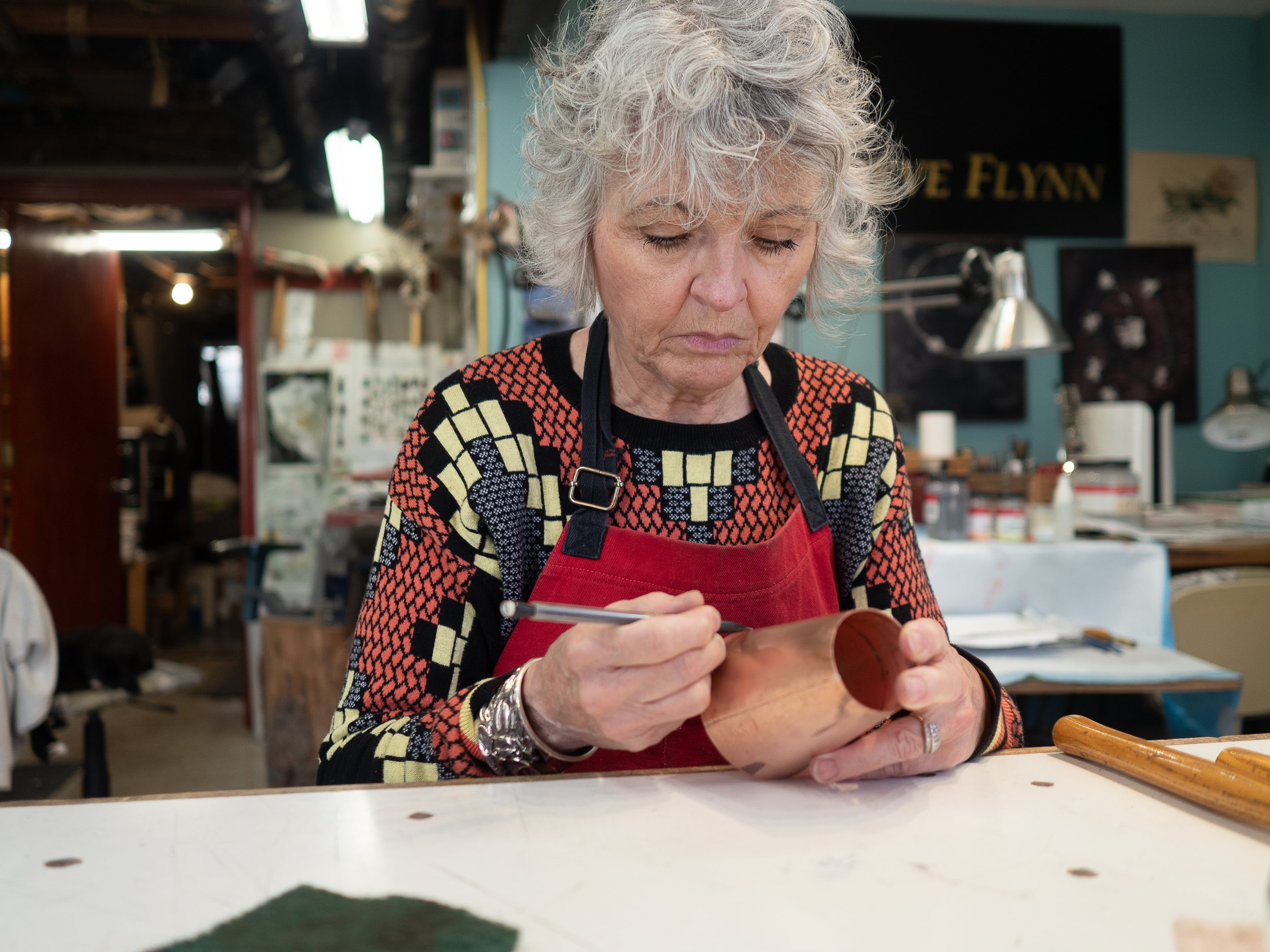 An elderly woman with curly gray hair wearing a colorful patterned sweater and red apron, working carefully on a crafting project in a workshop.