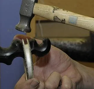 Close-up of a hand holding a hammer, with a tool in the hand, in a workshop setting.