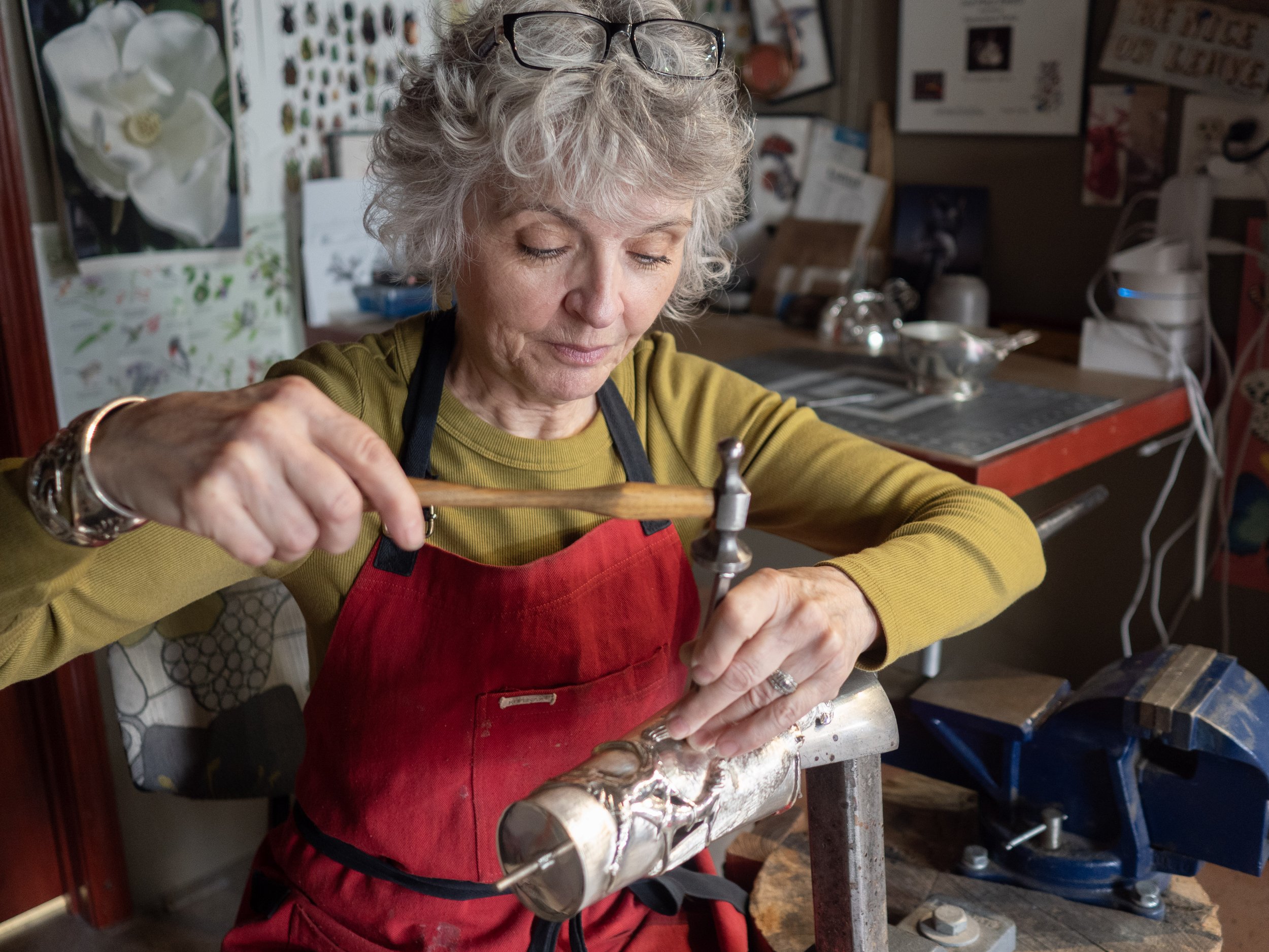Genevieve working on a metal project in a workshop surrounded by tools, artwork, and various objects.
