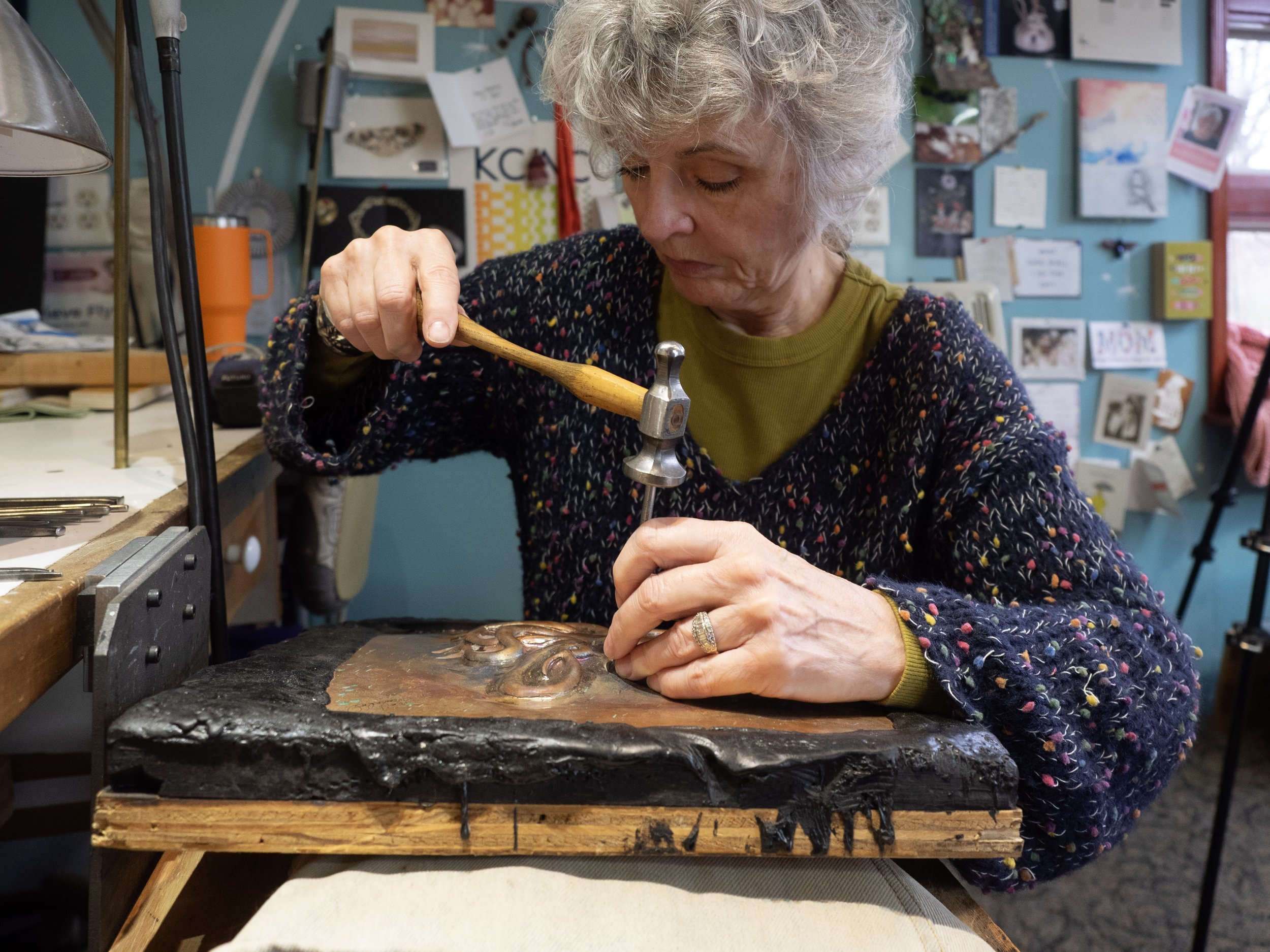 Genevieve working on a metal craft project at a cluttered workspace, holding a small hammer and working on a sheet of metal with various tools and artworks hanging on the wall behind her.