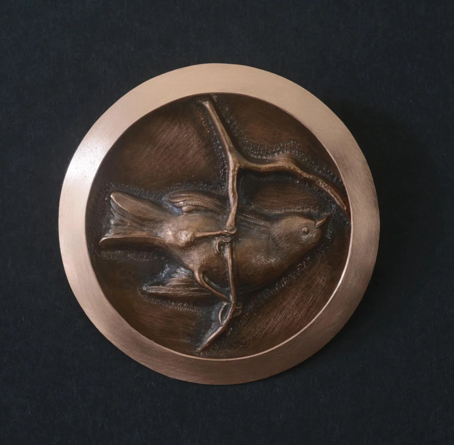 Round copper plaque with a raised design of a hummingbird perched on a curved branch.