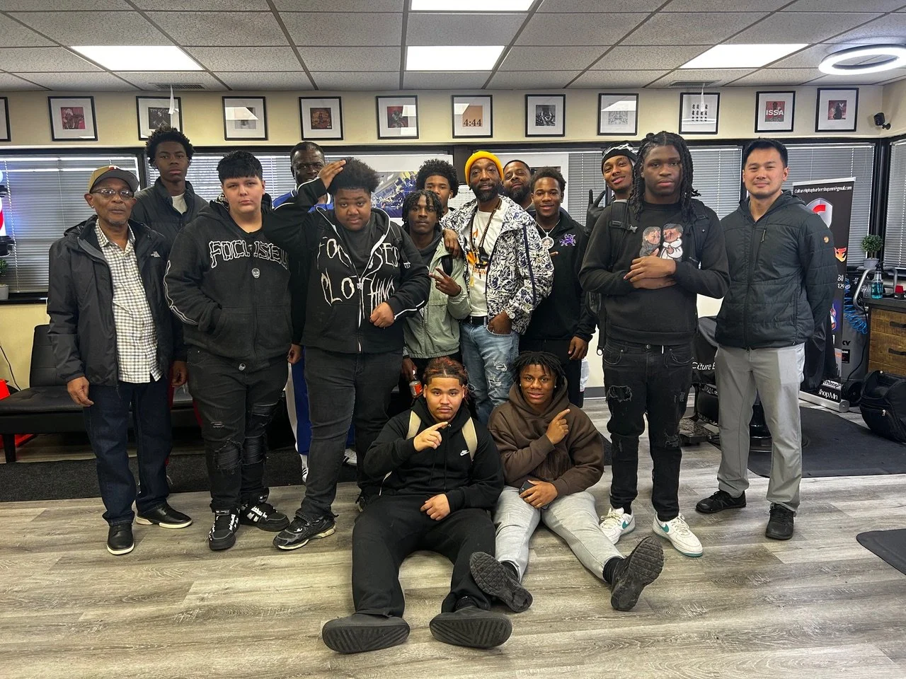 Group of fifteen young men and boys posing indoors in a room with framed pictures on the wall and windows with blinds. They are standing and sitting on the floor, some making hand gestures and smiling.