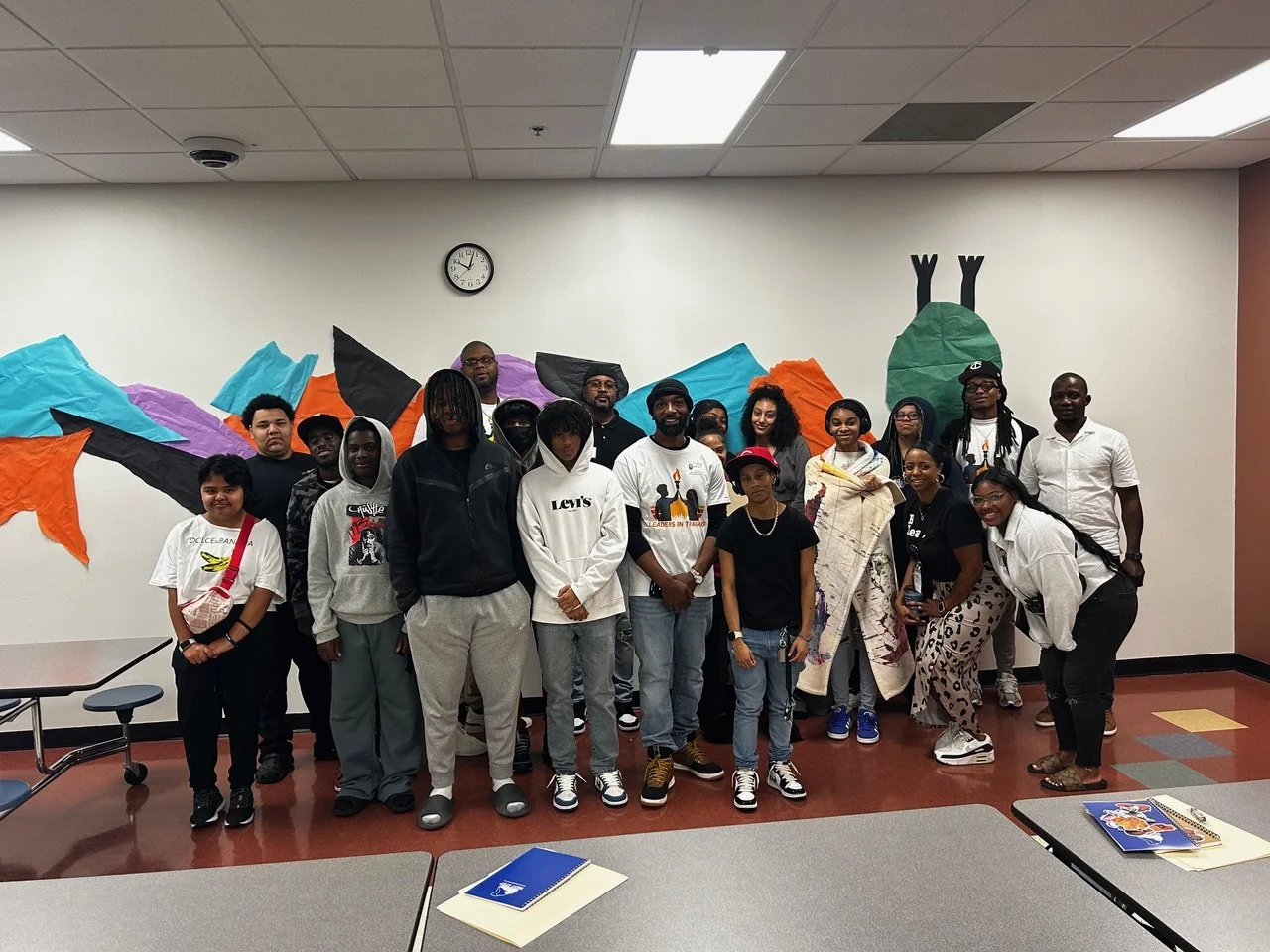A diverse group of people poses for a photo in a room with colorful paper mountain decorations and a large paper caterpillar on the wall. Some individuals are smiling, wearing casual clothes, and standing close together.