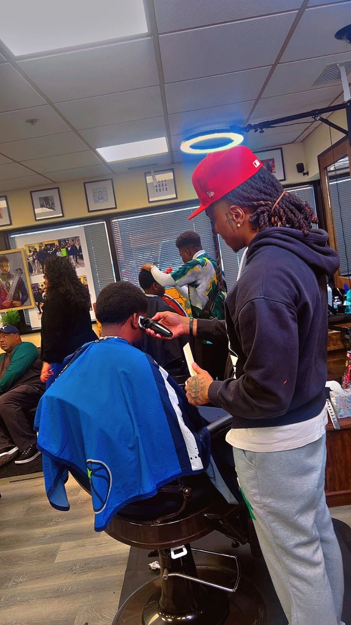 A man getting a haircut at a barbershop, with a barber holding clippers and a mirror, while other customers and barbers work in the background.