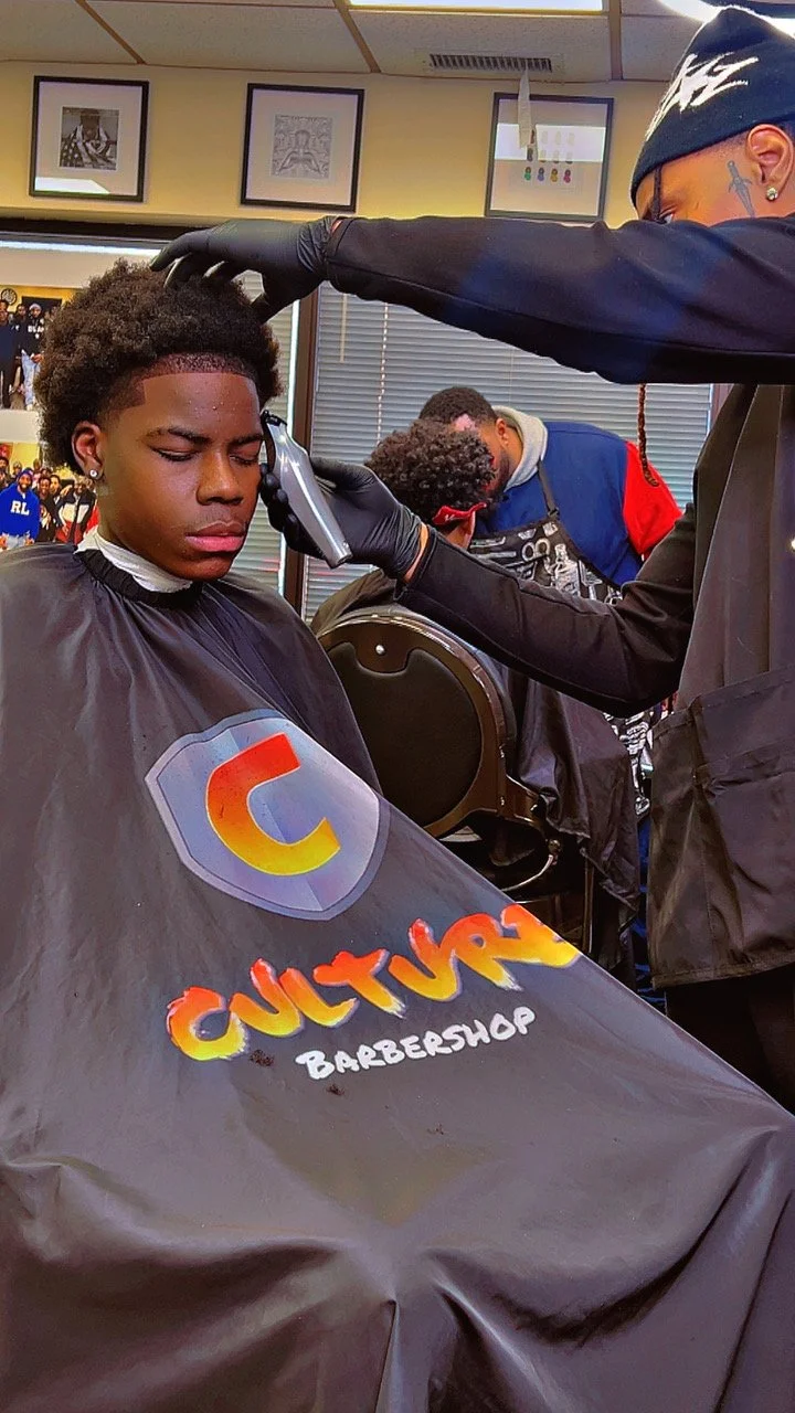 A young man getting a haircut at a barbershop, with a barber trimming his hair using clippers.