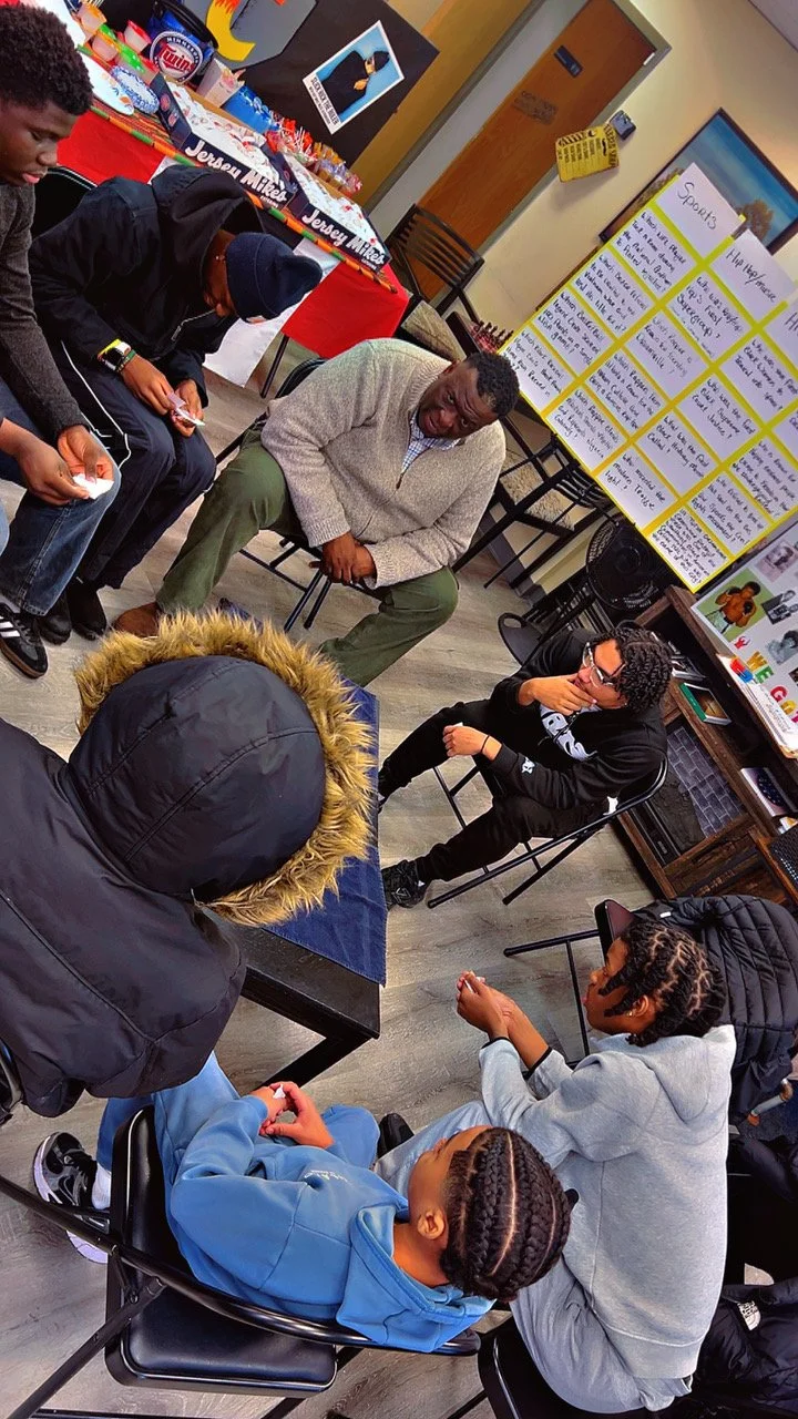 A group of young people sitting in a circle having a discussion in a classroom or community center. There is a person leading the discussion, and a colorful display or posters in the background.