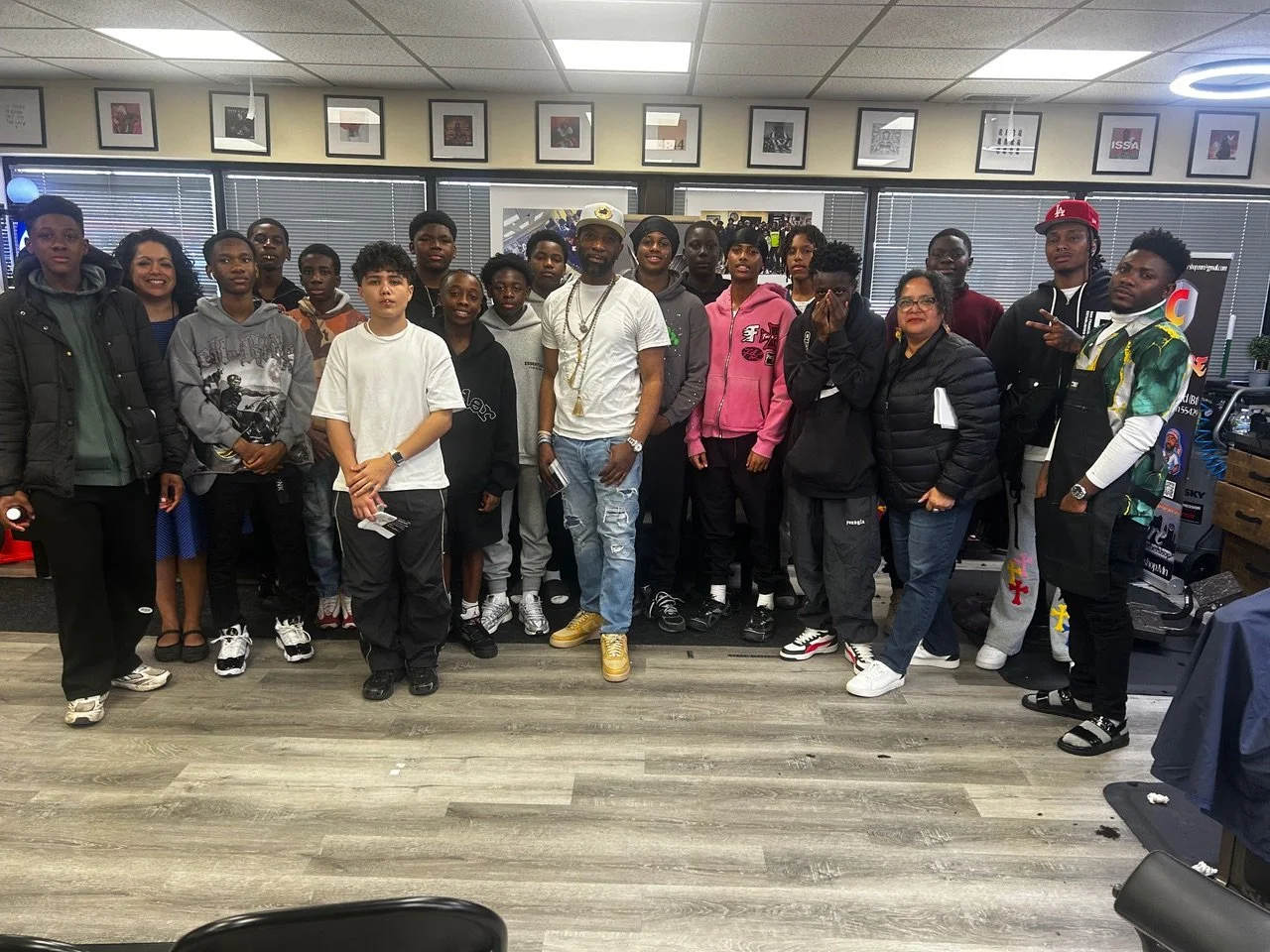 Group of people, including children and adults, gathered indoors for a photo, standing shoulder to shoulder, some smiling and others with neutral expressions, in a room with framed pictures on the wall and large windows with blinds.