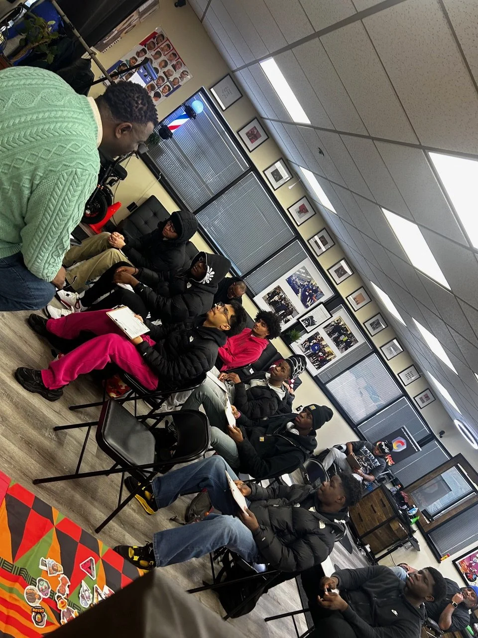 A group of young people sitting in a room, some holding notebooks, engaged in a discussion or presentation. There are framed pictures on the wall and a television displaying a barber pole.