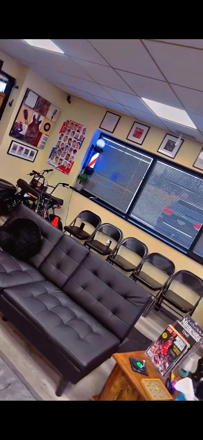 Indoor room with black leather sofa, four metal chairs aligned against the wall, a bicycle, framed pictures and posters, a barbershop pole, and a small wooden table with magazines and a wireless charger.