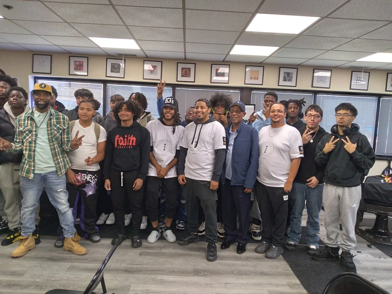 A diverse group of young men and a few older men posing for a photo in an indoor room with framed pictures on the wall and blinds on the windows.