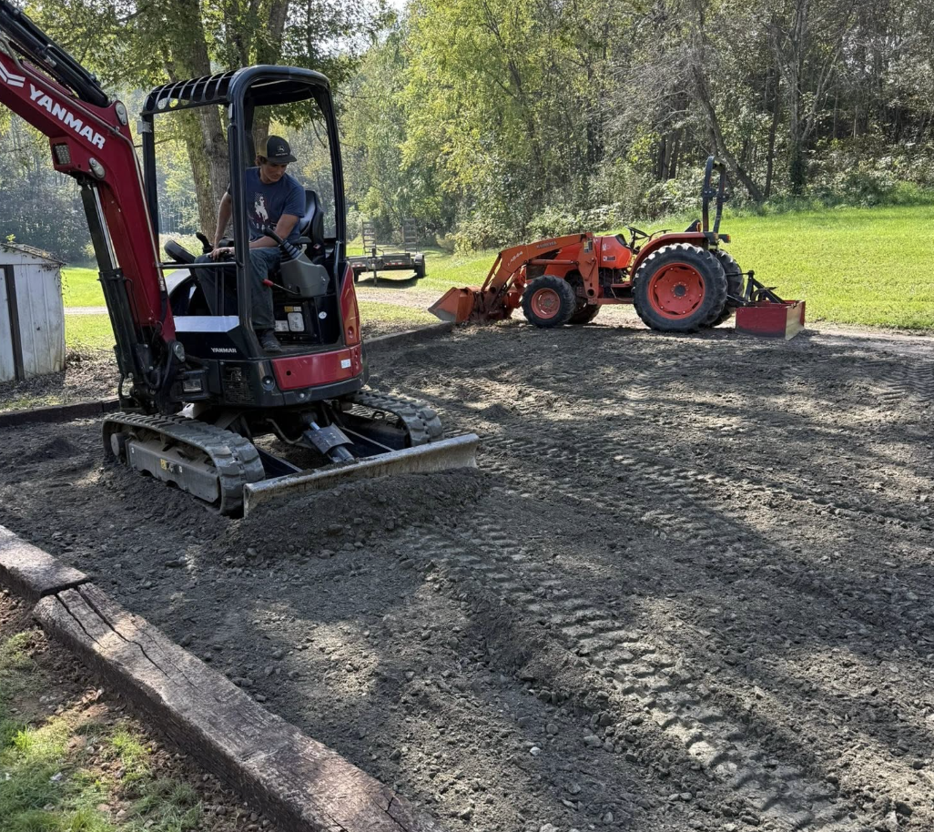 A person operating a small Yanmar excavator on a dirt patch with a red tractor in the background, surrounded by trees and green grass.