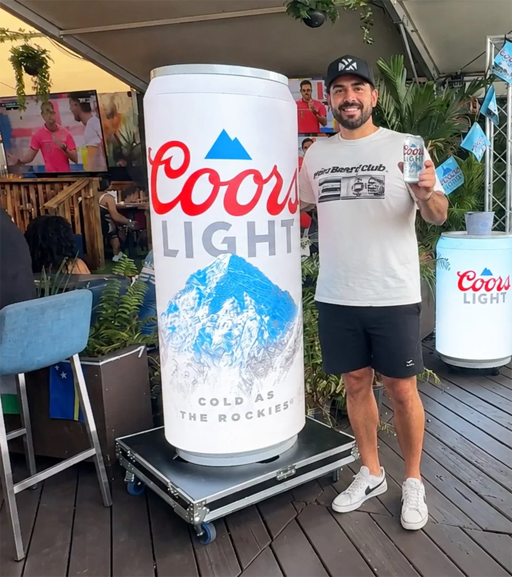 A man holding a Coors Light beer can next to the MotionCan Tall Can LED Display. He is smiling and wearing a white t-shirt, black shorts, and white sneakers at a World Cup 2026 event, with the supporting MotionCan Refrigerated can in the background.