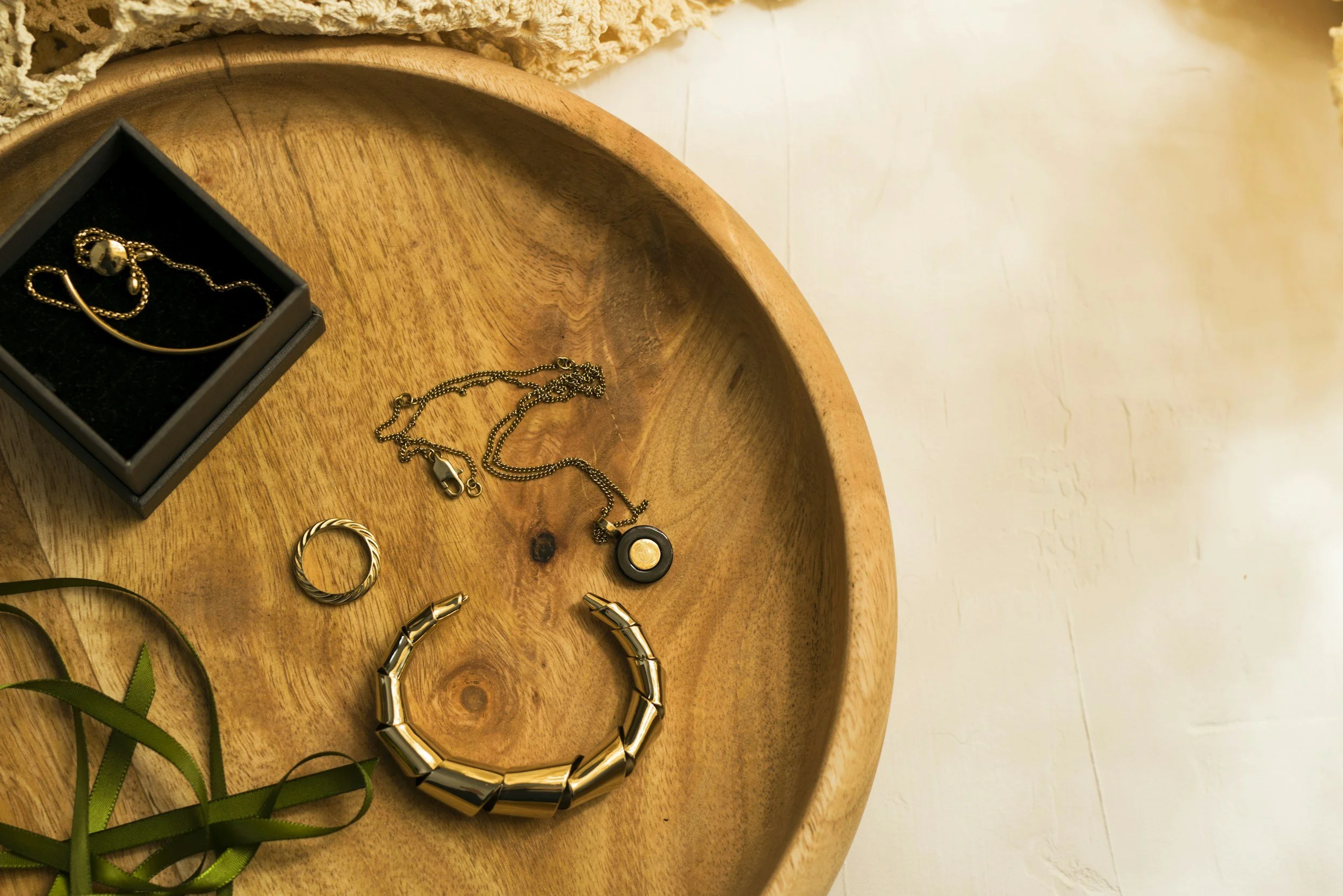 A round wooden tray with various pieces of gold jewelry, including a chain necklace, a bangle, and a ring, along with a small black jewelry box containing a bracelet, on top of a light-colored surface.