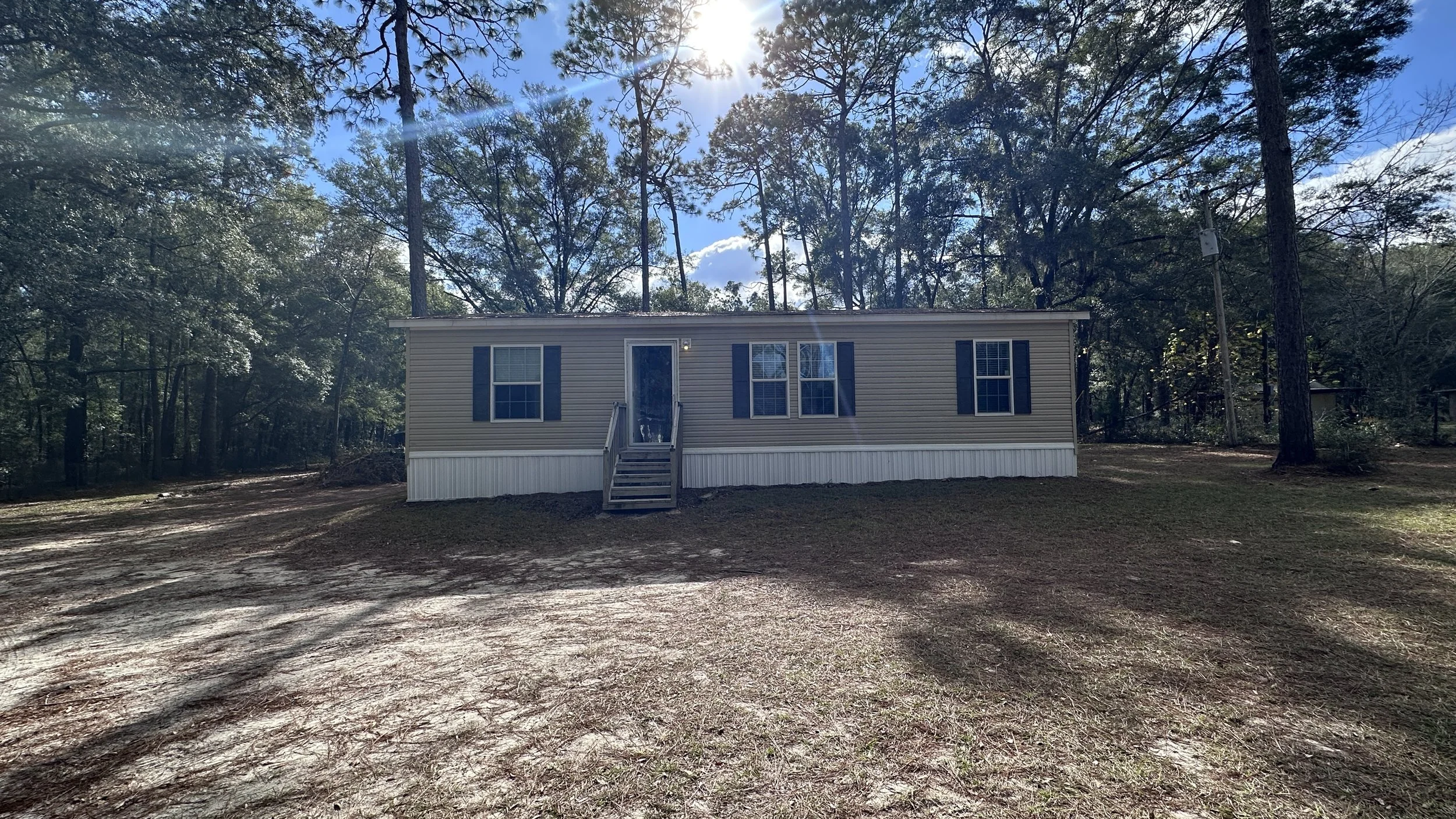 A beige mobile home with four windows and a door, surrounded by trees and a dirt yard under sunlight.