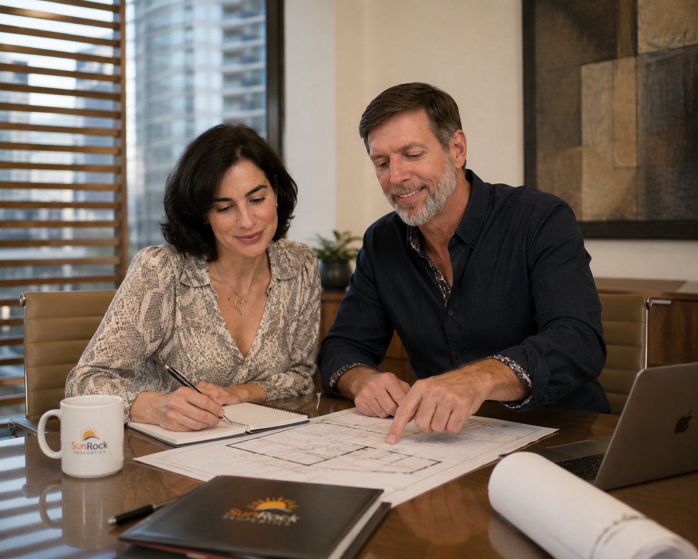 Two people, a woman with dark hair and a man with gray hair and beard, sit at a table reviewing architectural plans. They are in an office with large windows and wooden blinds. There is a labeled coffee mug, a laptop, a notebook, and rolled-up blueprints on the table.