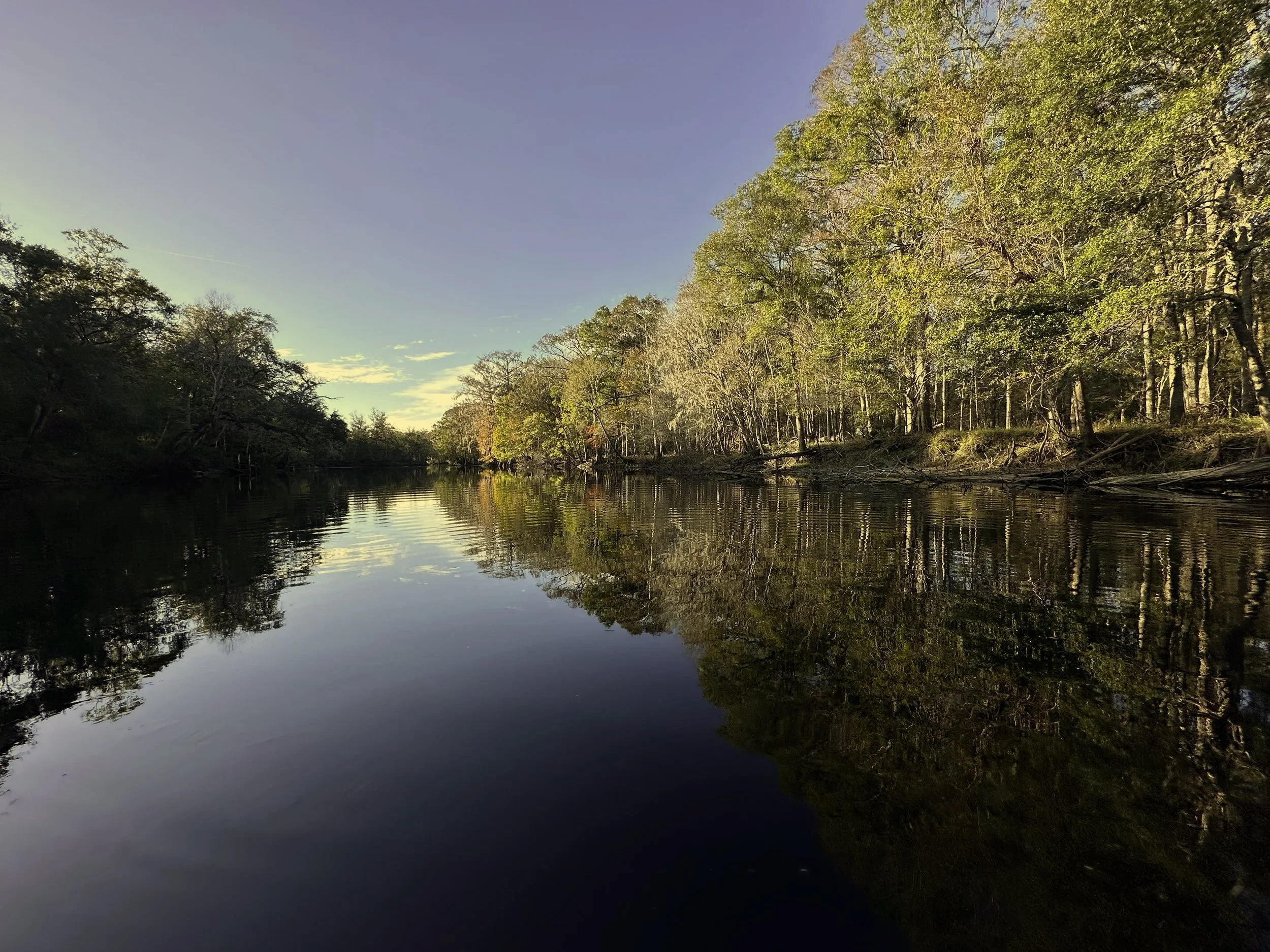 A peaceful river scene with calm water reflecting the trees on the right bank and the sky on the left. The sky is partly cloudy with a gradient from blue to light yellow near the horizon. The trees are lush and green, indicating late spring or early summer.