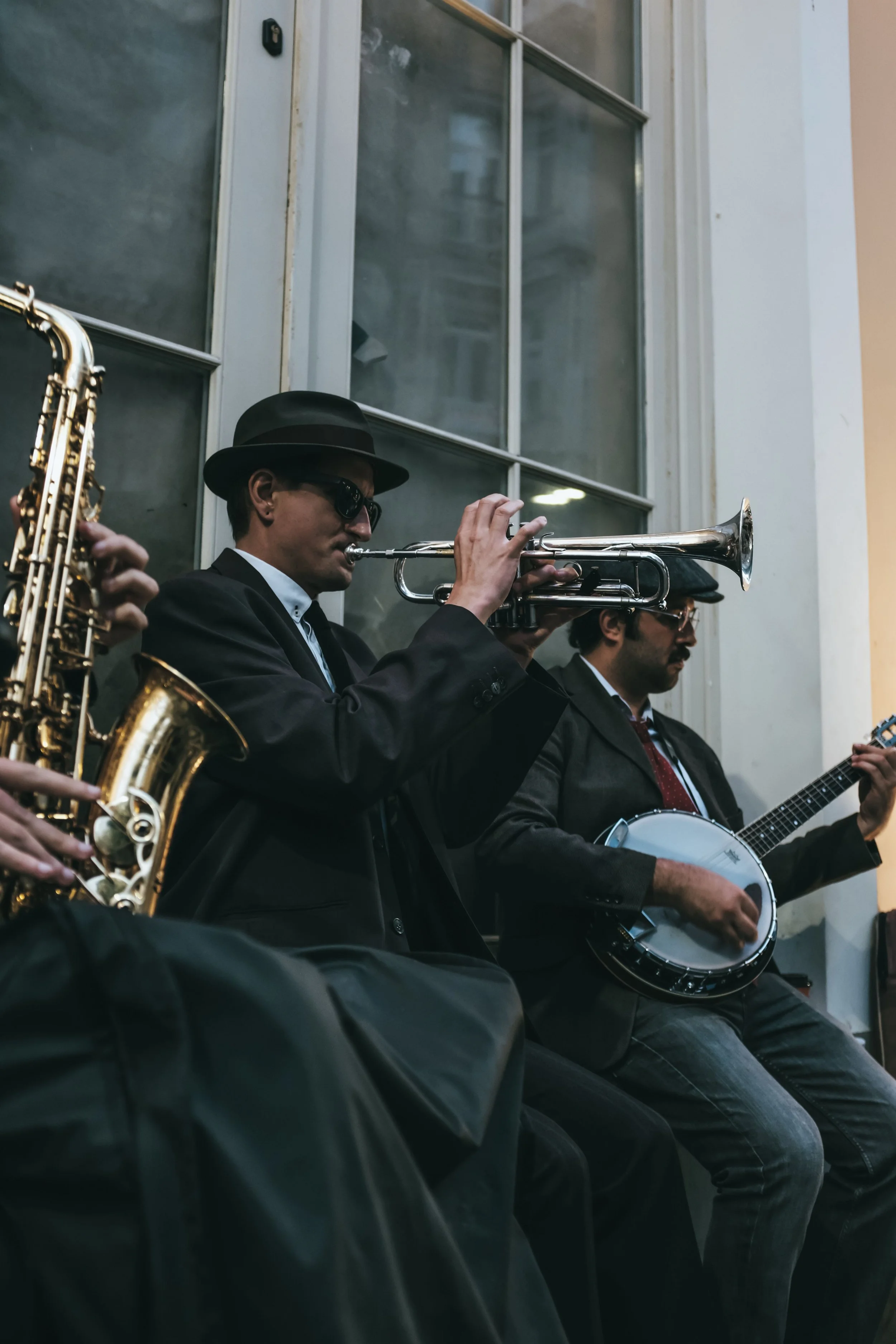 Two men dressed in suits and sunglasses are playing musical instruments, one with a trumpet and the other with a banjo, sitting against a windowed wall.