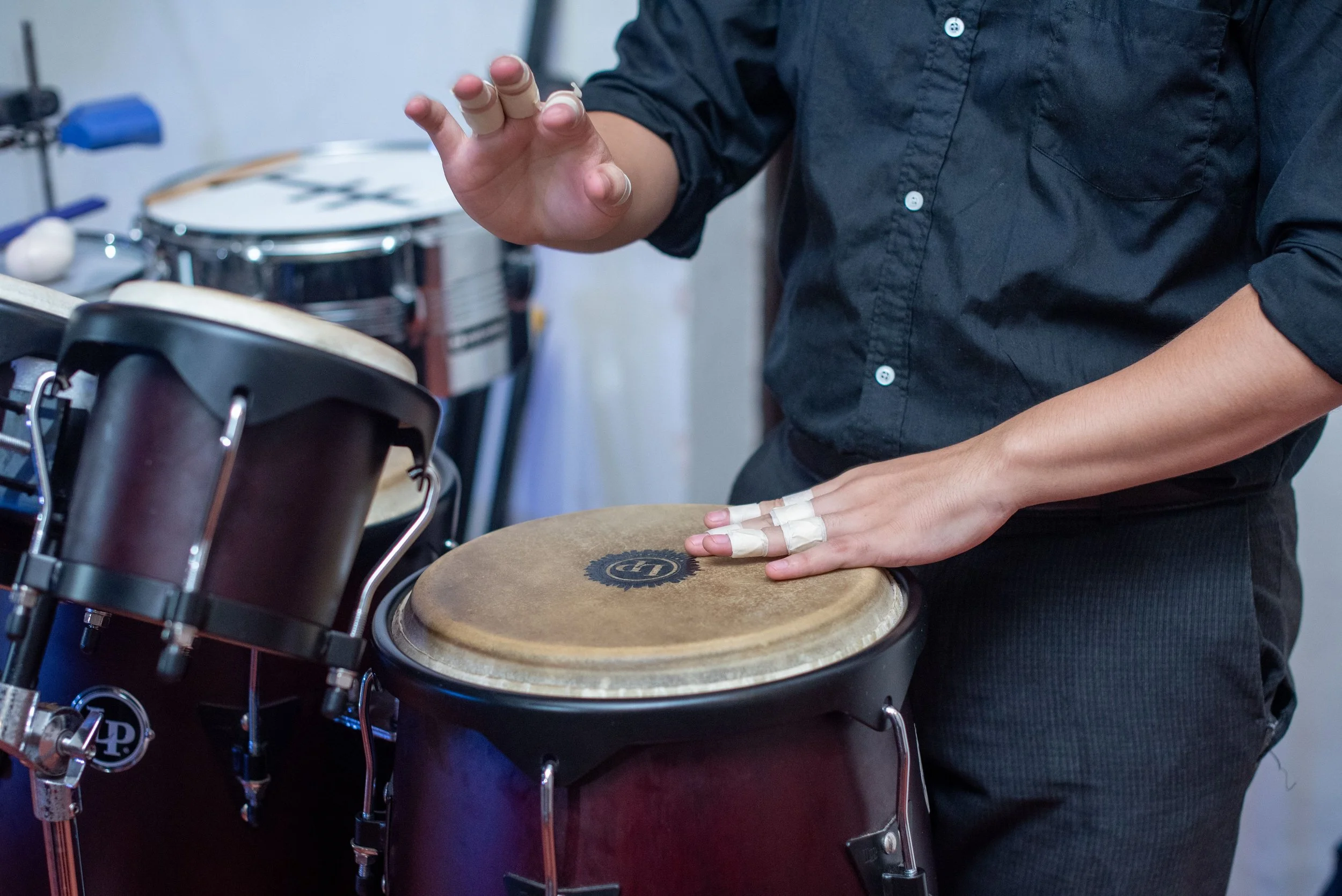Person playing bongos with finger tapping on drumhead, various drums and percussion instruments in background.