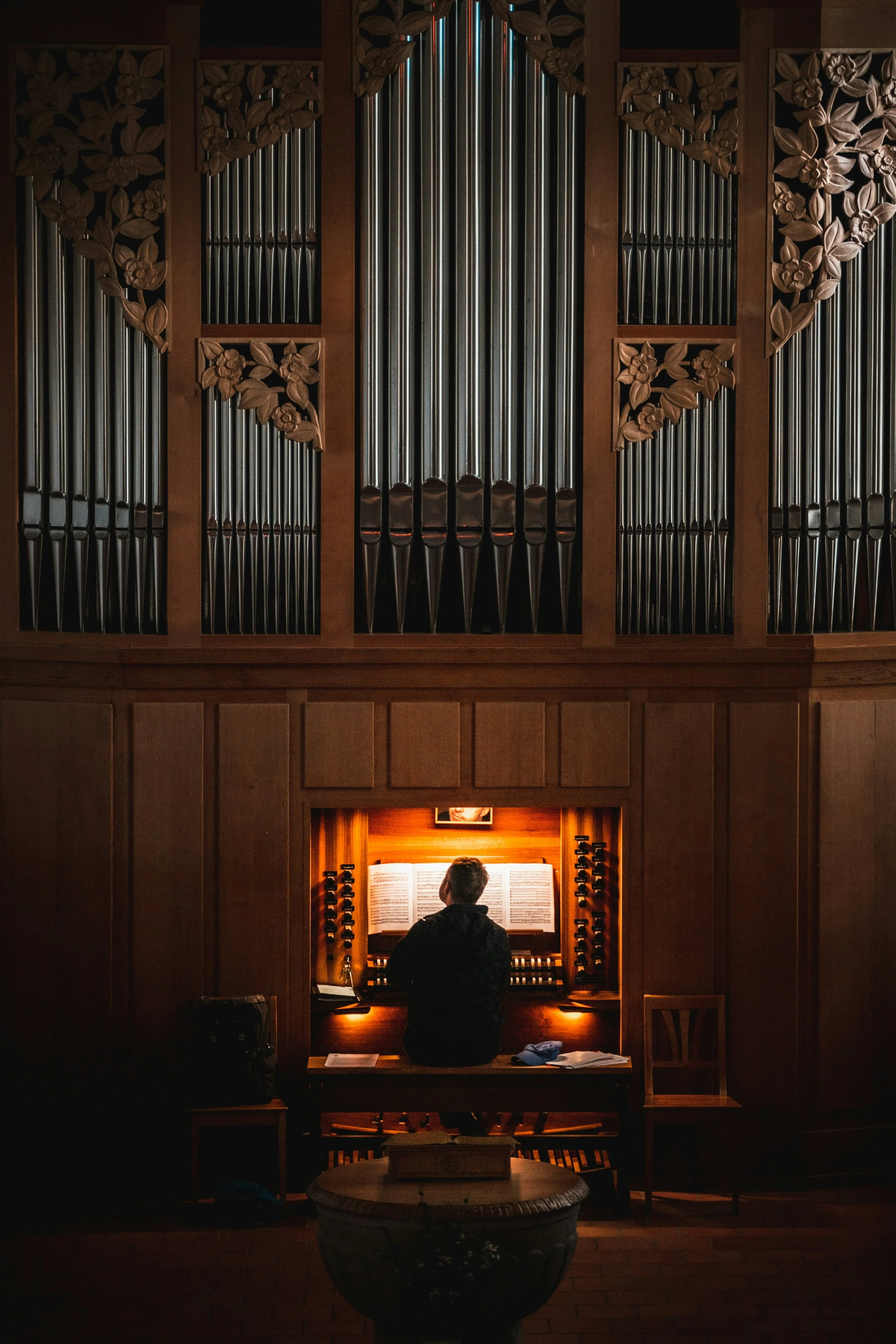 A person sitting at an organ console in a dimly lit room, with intricate wooden carvings and pipes above, and a small framed picture on the wall behind.