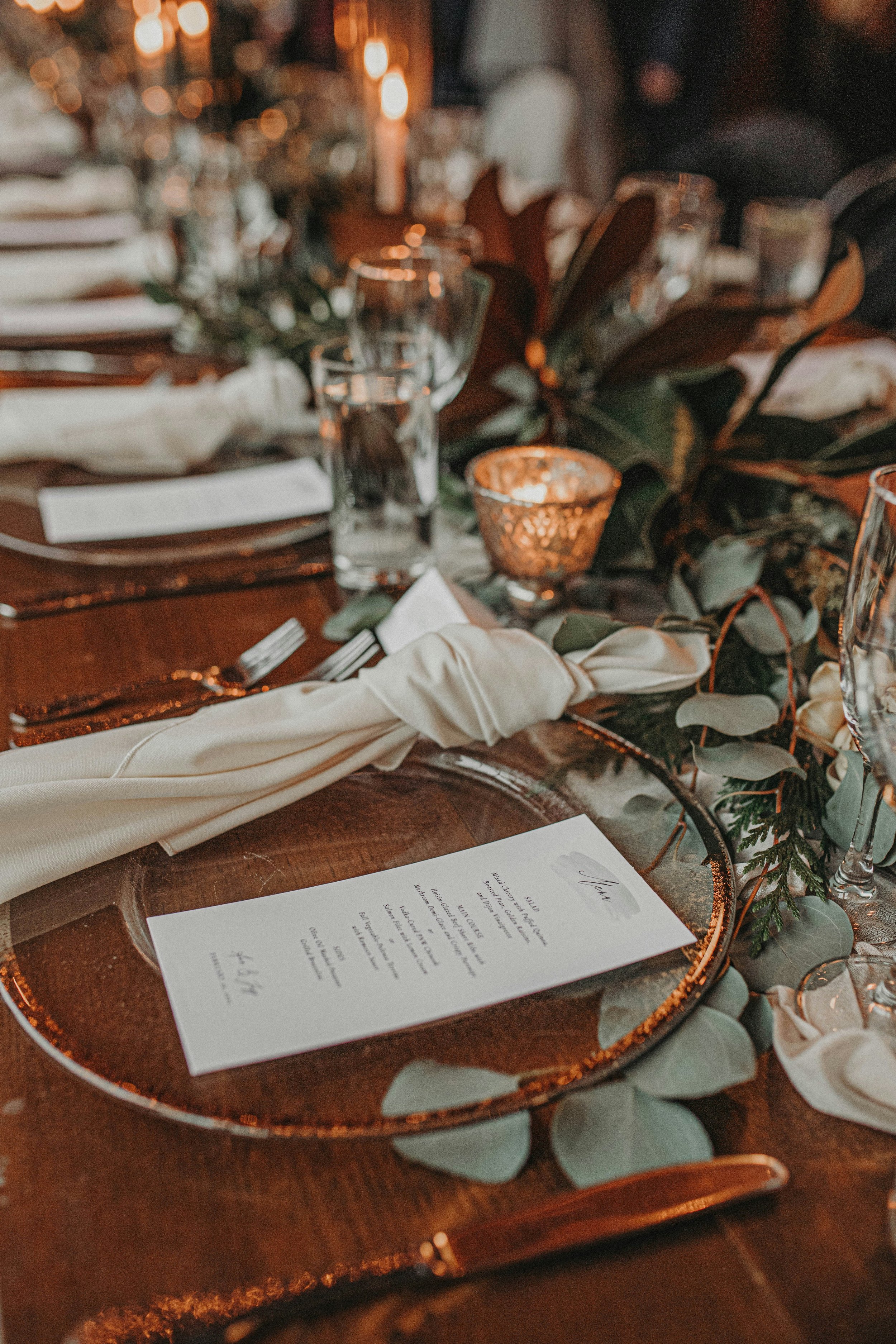 A table set for a formal event with a menu, glassware, napkins, and greenery in a warm, dimly lit environment.