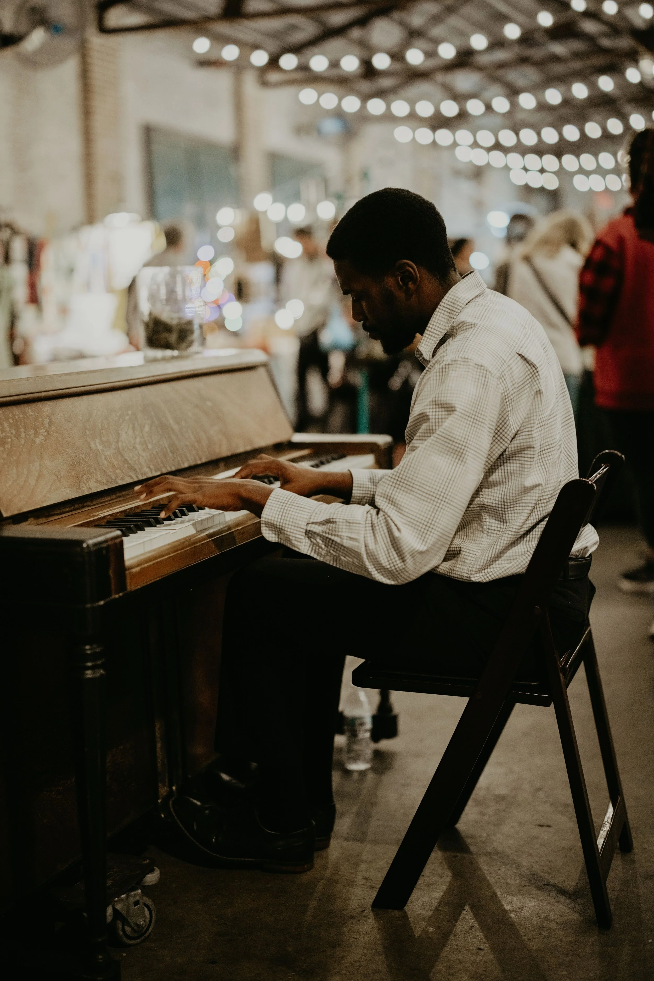 Man playing piano in a lively indoor setting with string lights and people walking around in the background.