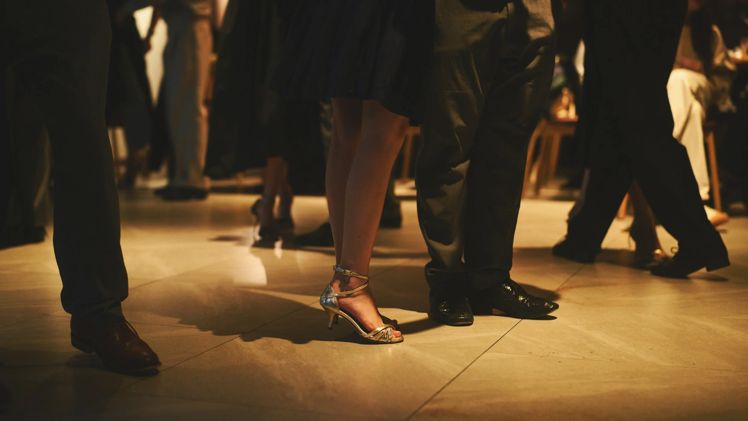 People dancing closely together in a dimly lit room, focusing on the lower half of their bodies, with women wearing high-heeled shoes and men in dark pants.