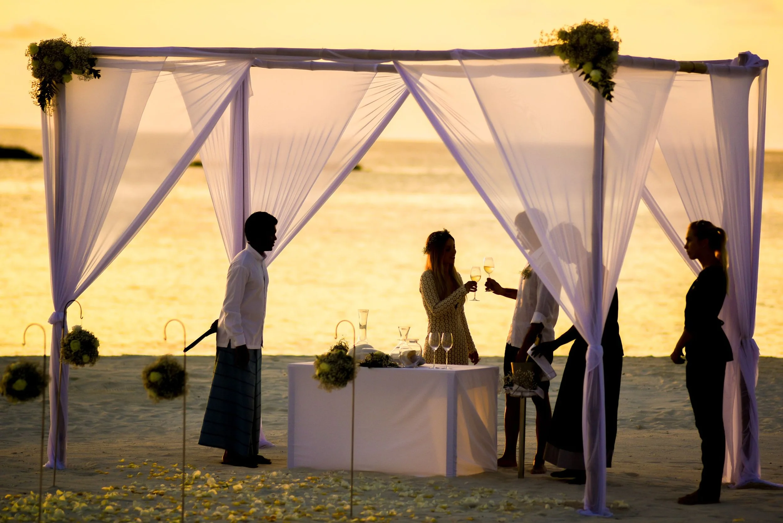A wedding ceremony setup on the beach during sunset with draped white fabric and floral arrangements, with a couple exchanging vows and guests standing nearby.