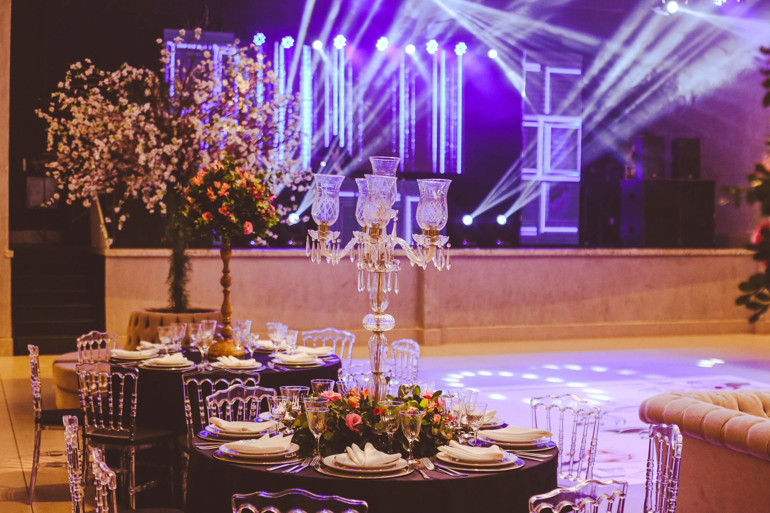 Elegant event setup with a large round table decorated with floral centerpieces, fine glassware, and white napkins, surrounded by transparent ghost chairs. In the background, a stage with vibrant purple lighting, spotlights, and a floral arrangement adds to the festive atmosphere.