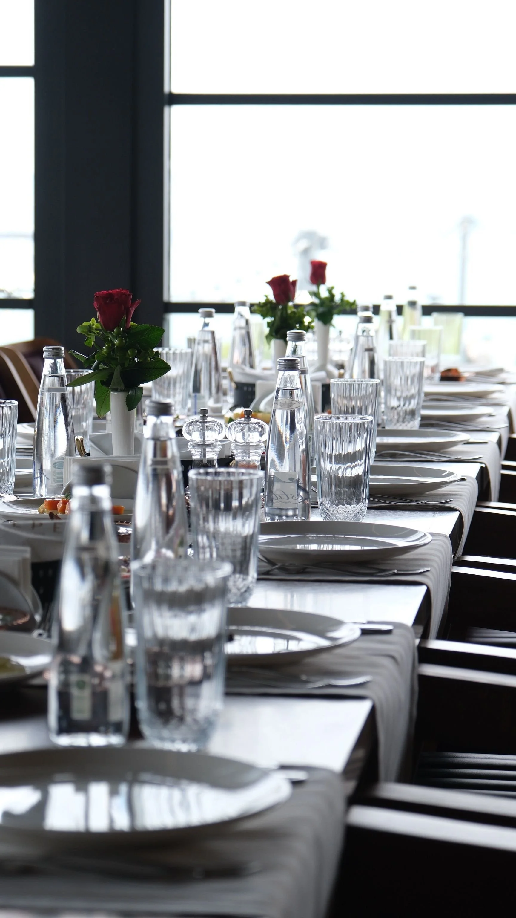 A dining table set for a formal event with plates, silverware, glasses, and floral centerpieces, near large windows letting in natural light.