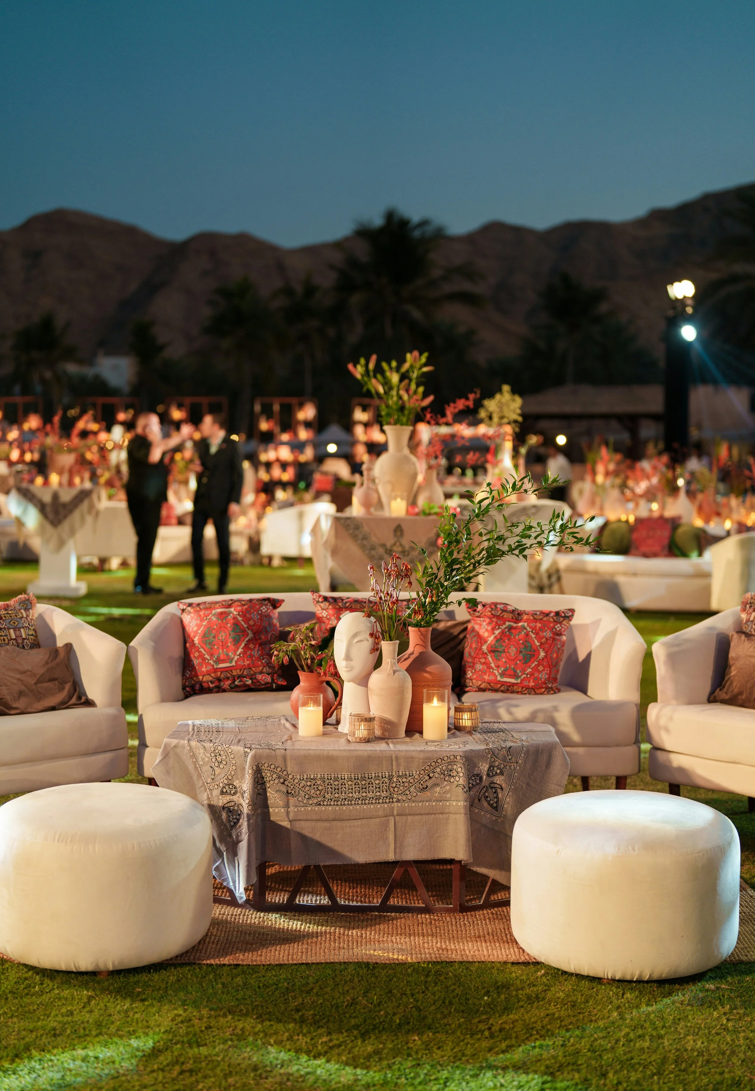 Outdoor evening event with a cozy seating area, decorated with vases, candles, and pillows, with tables and lights in the background, mountains, and palm trees.