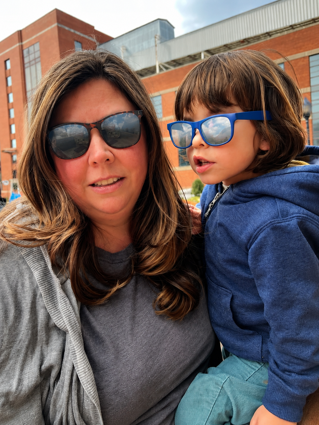 A woman and a young boy with sunglasses outdoors, with a brick building in the background.