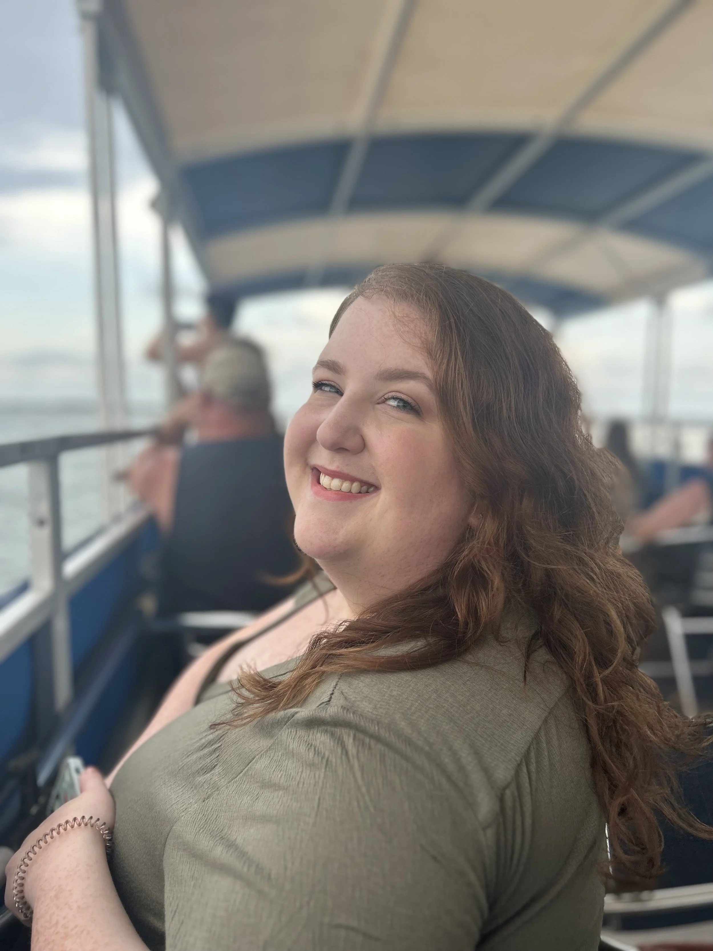 A woman with long, wavy, light brown hair and a broad smile, sitting on a boat with a blue striped canopy, with other people in the background.